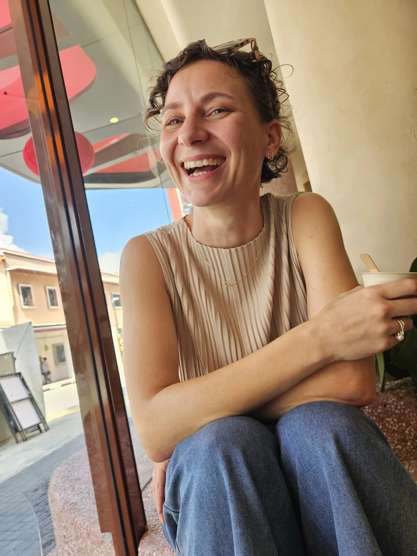 A woman with short curly hair and glasses resting on her head, smiling while sitting inside a café near a window, holding a cup.