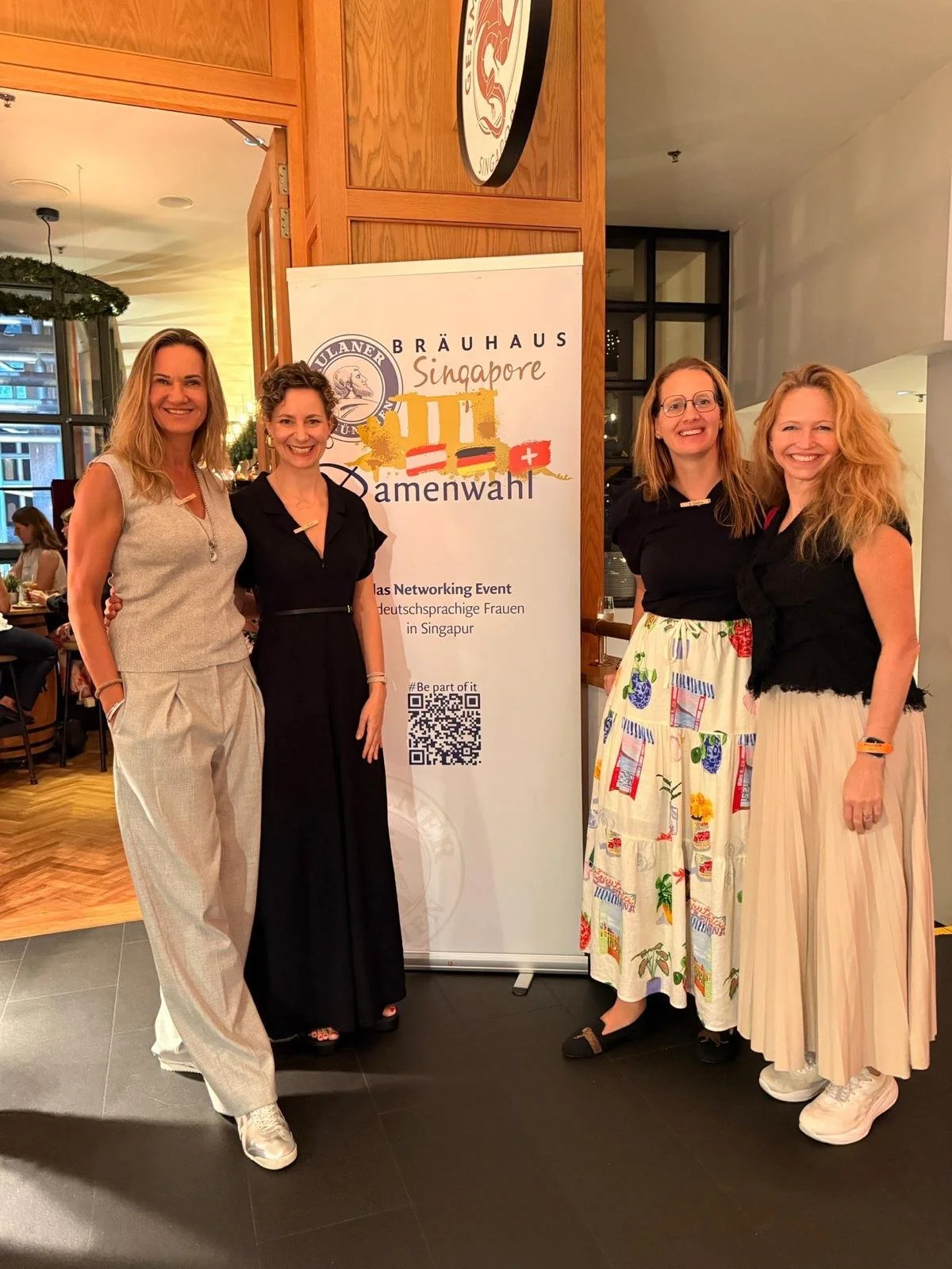 Four women standing together indoors near a sign for a networking event for German-speaking women in Singapore. They are smiling and dressed in casual summer attire.