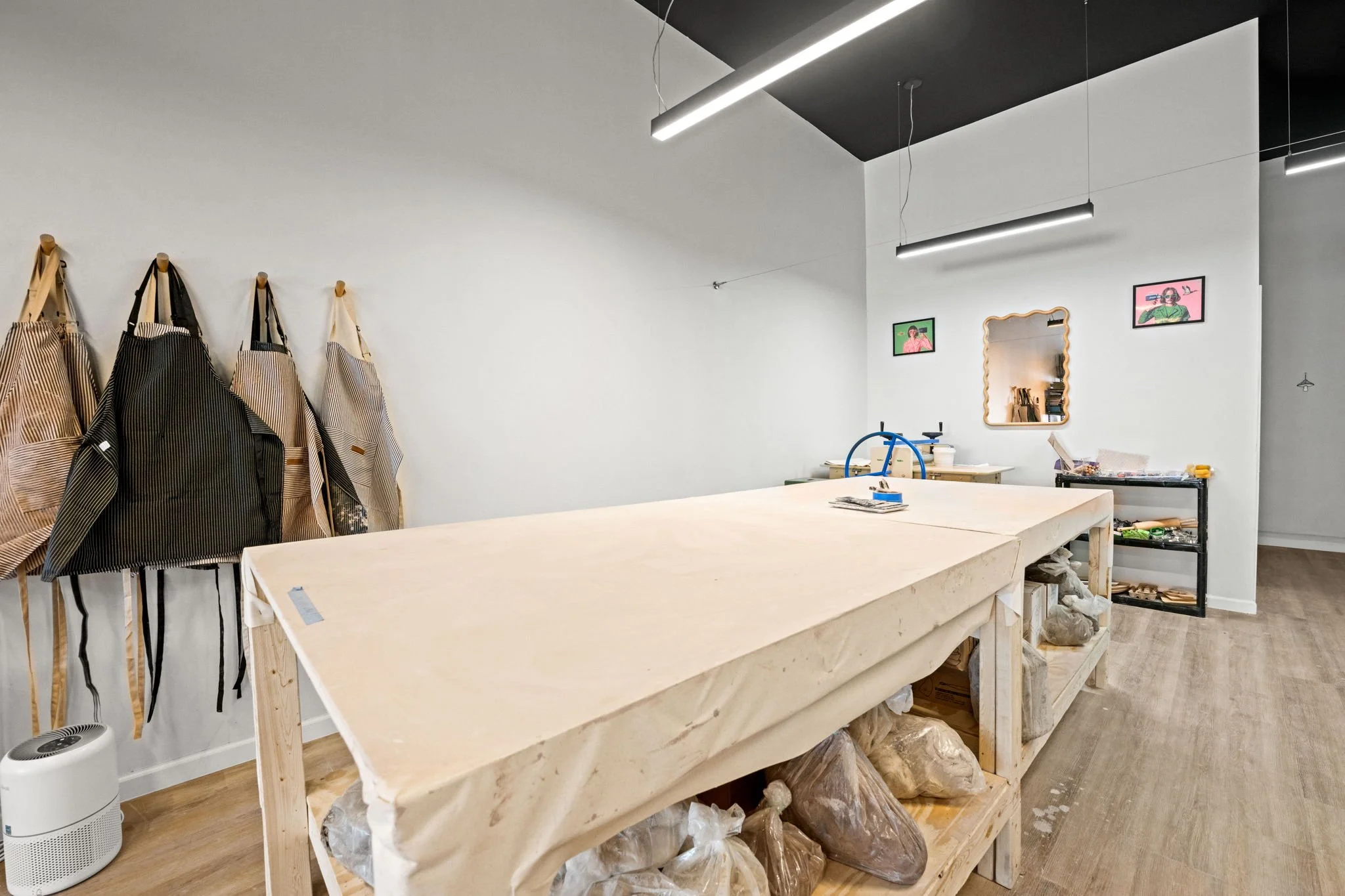 Interior of a boutique or workshop with fabric aprons hanging on hooks, a large wooden work table, a black shelving unit with tools, framed artwork on the wall, and a mirror.