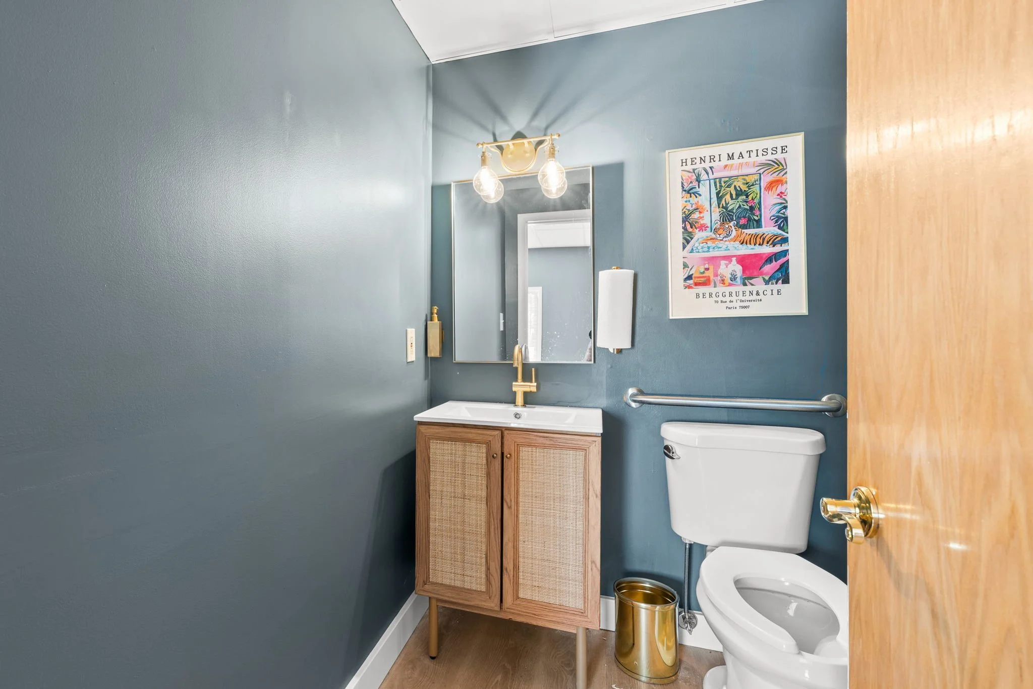 Small bathroom with blue walls, wooden cabinet with a white sink, mirror, gold faucet, white toilet, gold trash can, and colorful poster on the wall.