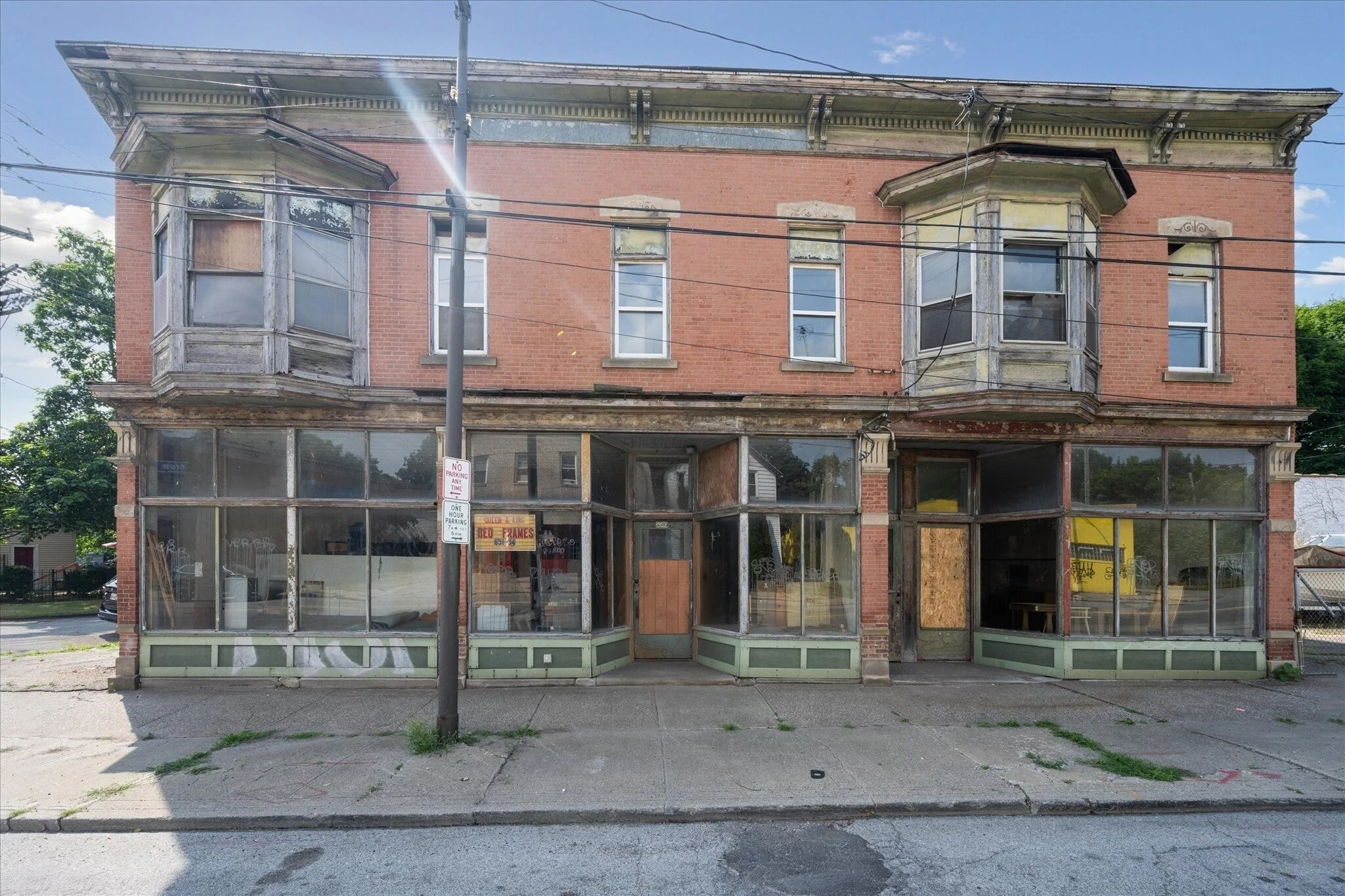 An abandoned, two-story brick building with boarded-up windows, some windows covered in graffiti, and a broken storefront with missing glass panes. The sidewalk in front has overgrown patches of grass and a streetlight pole.