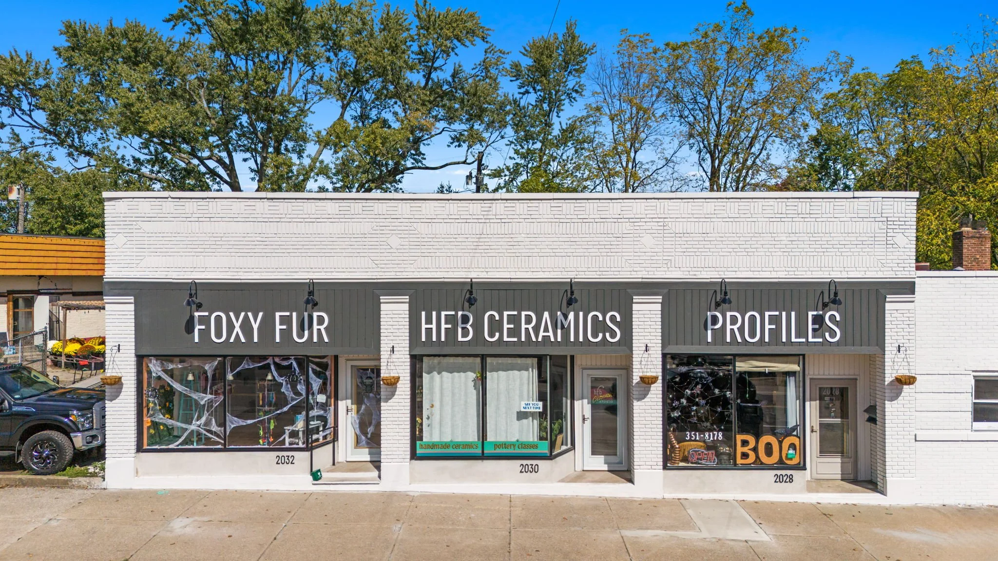 Storefront with three signs: Foxy Fur, HFB Ceramics, and Profiles, decorated with cobwebs, in a commercial area with trees in the background.
