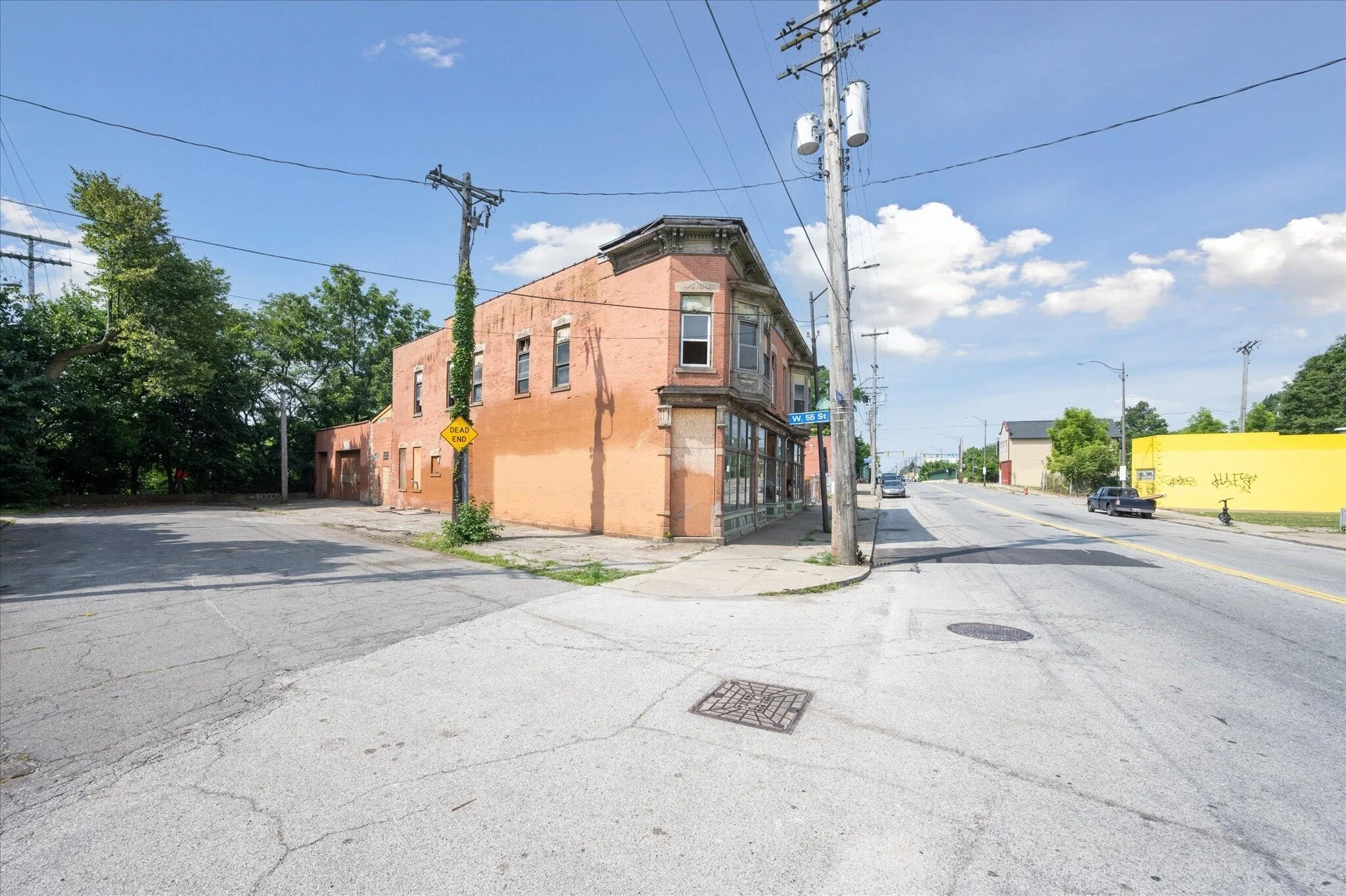 A street corner with a large, two-story brick building on the corner and a stop sign that says 'Dead End.' The street signs show 'W. 55th St.' The scene is sunny with a few clouds, and there are power lines and trees in the background.