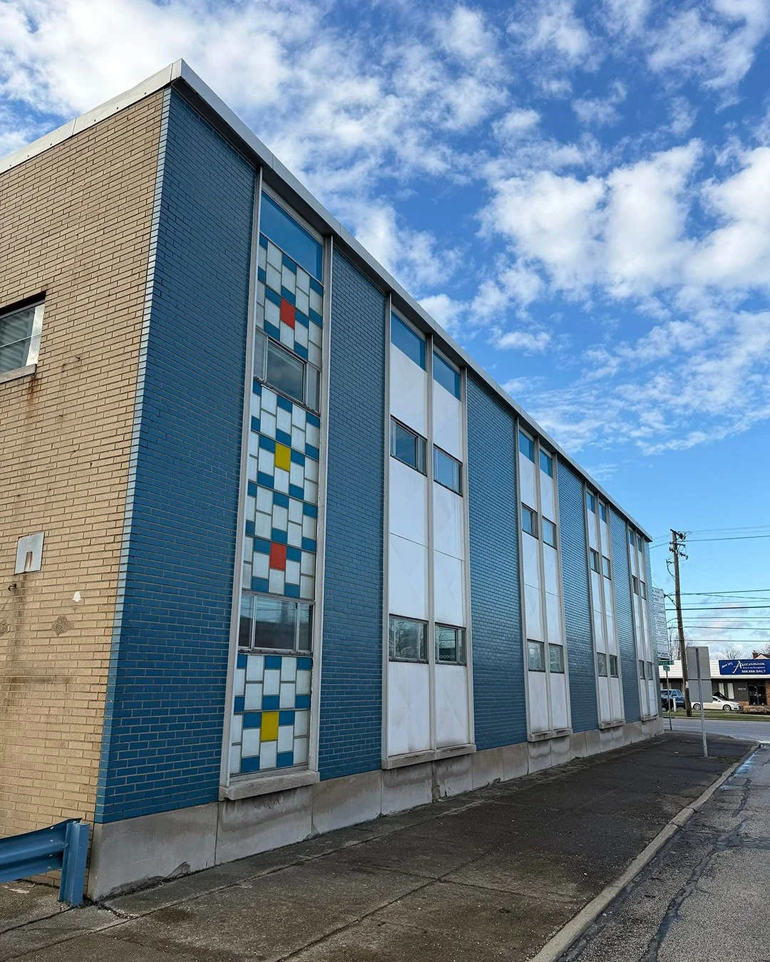 A multi-story building with blue and beige brick exterior, featuring vertical stained glass windows with geometric patterns, under a partly cloudy blue sky.
