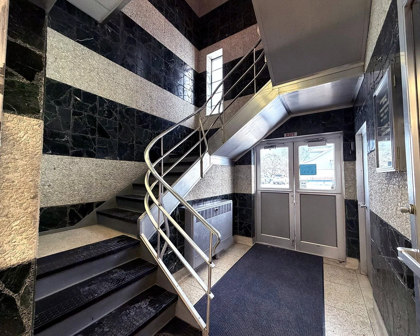 Interior of a building lobby with black and beige tiled walls, a staircase with black steps and a metal railing, a glass door exit, and a black rug on the beige tile floor.