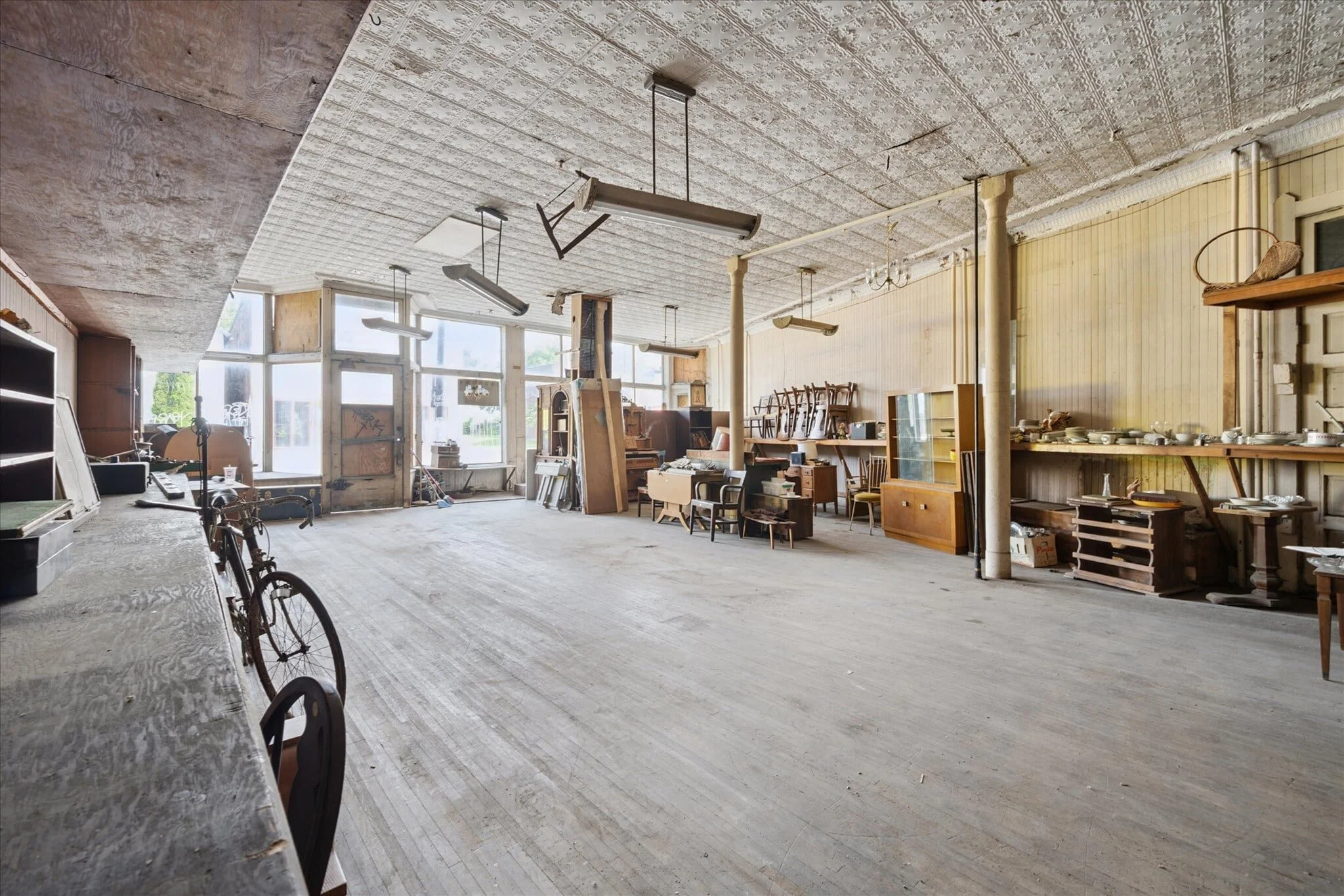 Empty interior of an old, abandoned store or workshop with various furniture and items scattered around, including chairs, cabinets, and a bicycle, with large windows and ceiling lights.