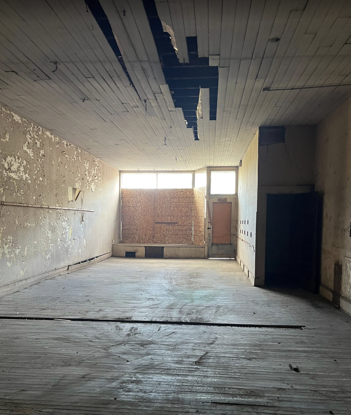 An empty, derelict room with peeling paint on the walls, boarded-up windows, and a damaged ceiling.