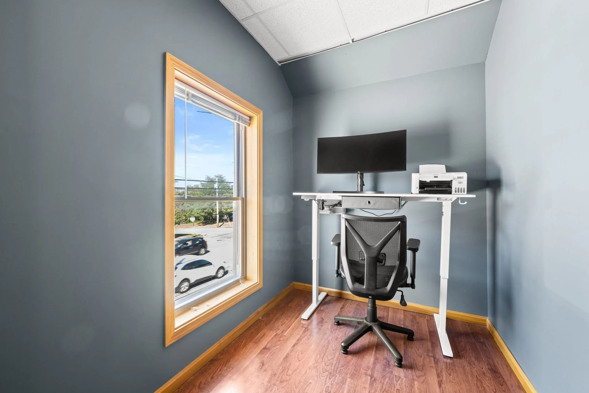 Small office with blue walls, wooden trim, window showing cars outside, black office chair, desk with black monitor, printer.