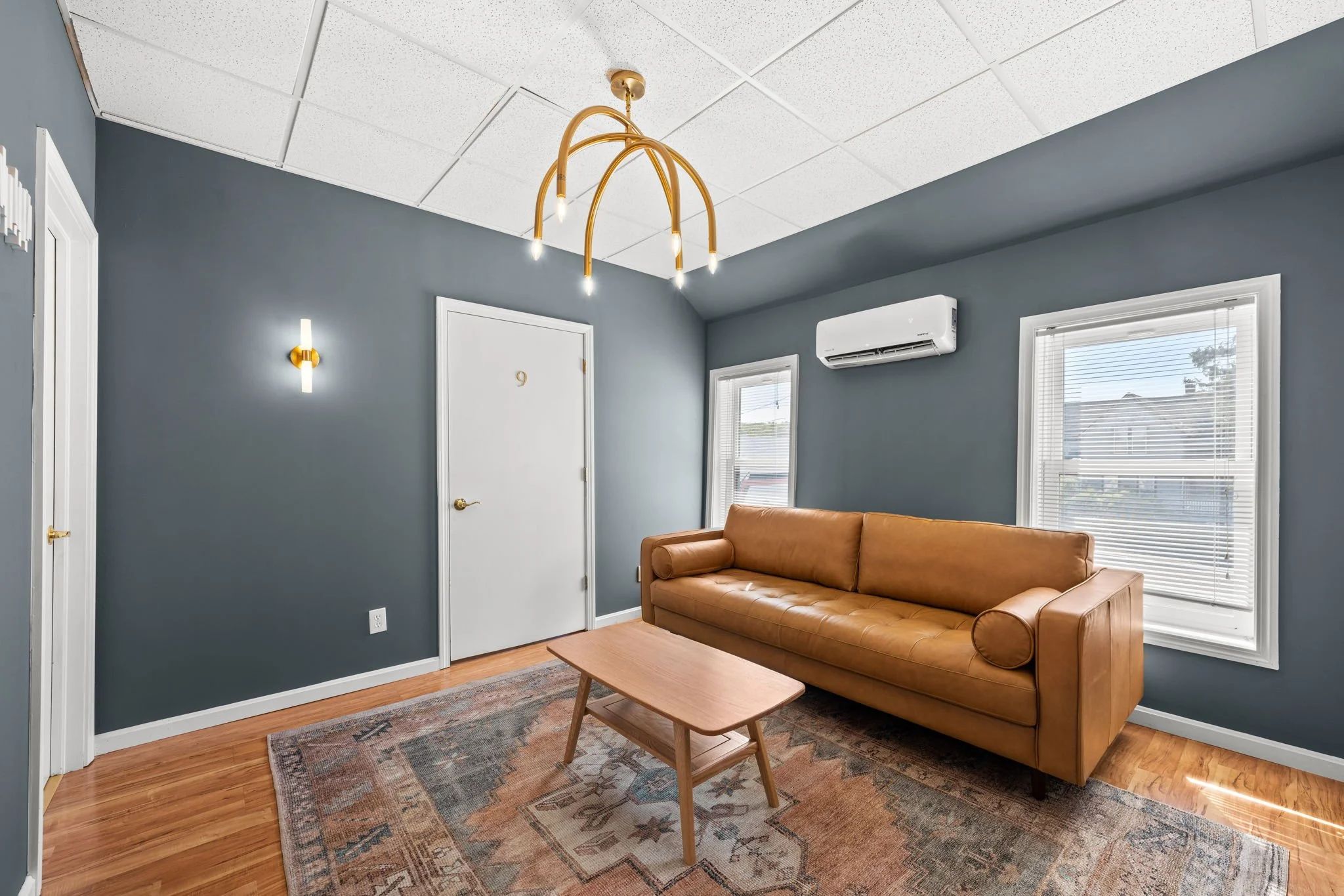 Living room with navy blue walls, hardwood floors, a tan leather sofa, a wooden coffee table, two windows with blinds, a wall-mounted air conditioning unit, a door labeled 9, a wall sconce, and a modern gold chandelier.