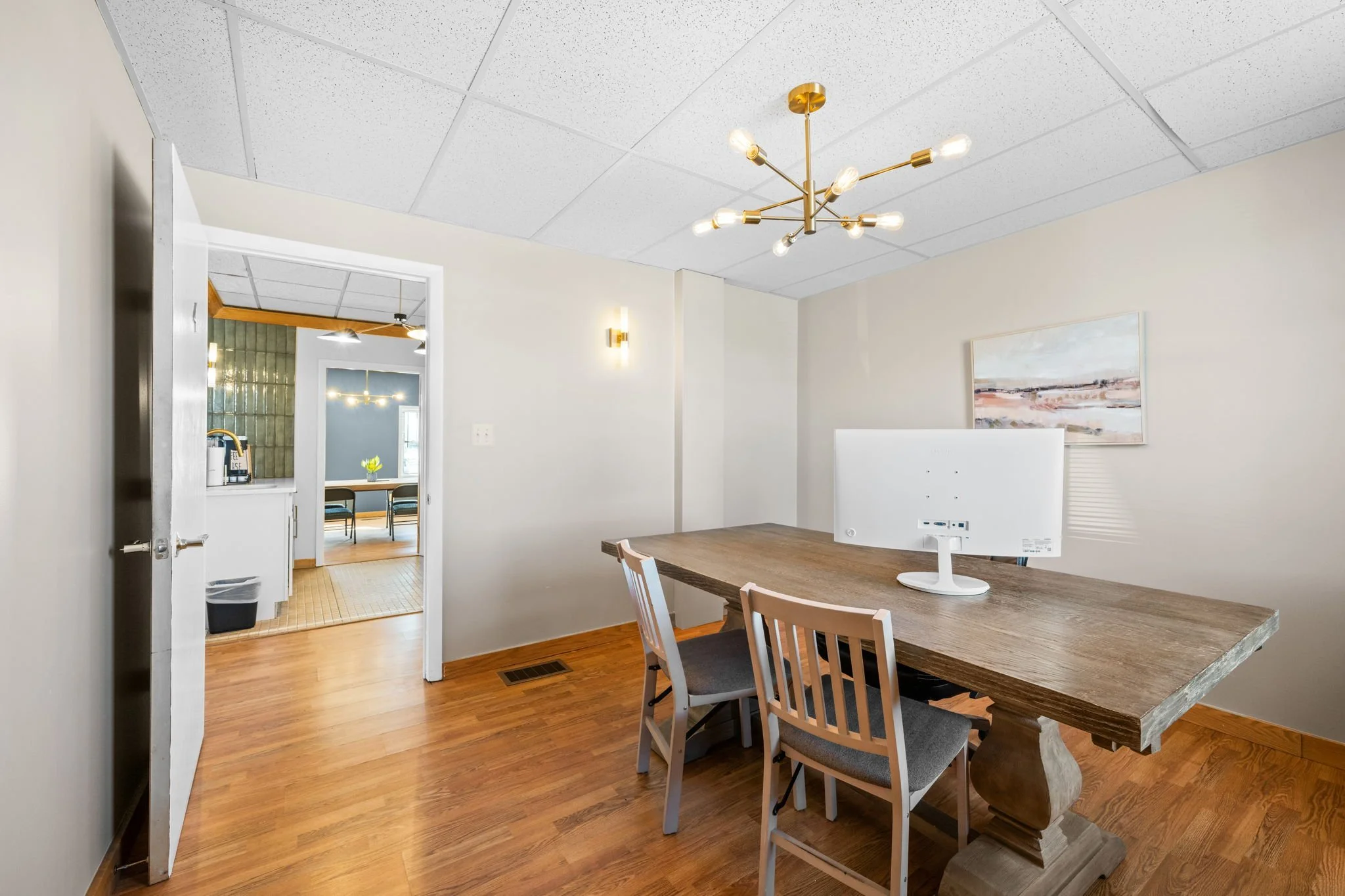 A dining area with a wooden table, four chairs, a white computer monitor, a modern gold chandelier, a wall-mounted light fixture, and a landscape painting. An open doorway shows a kitchen with a green tile wall and a room with a dining table and chai