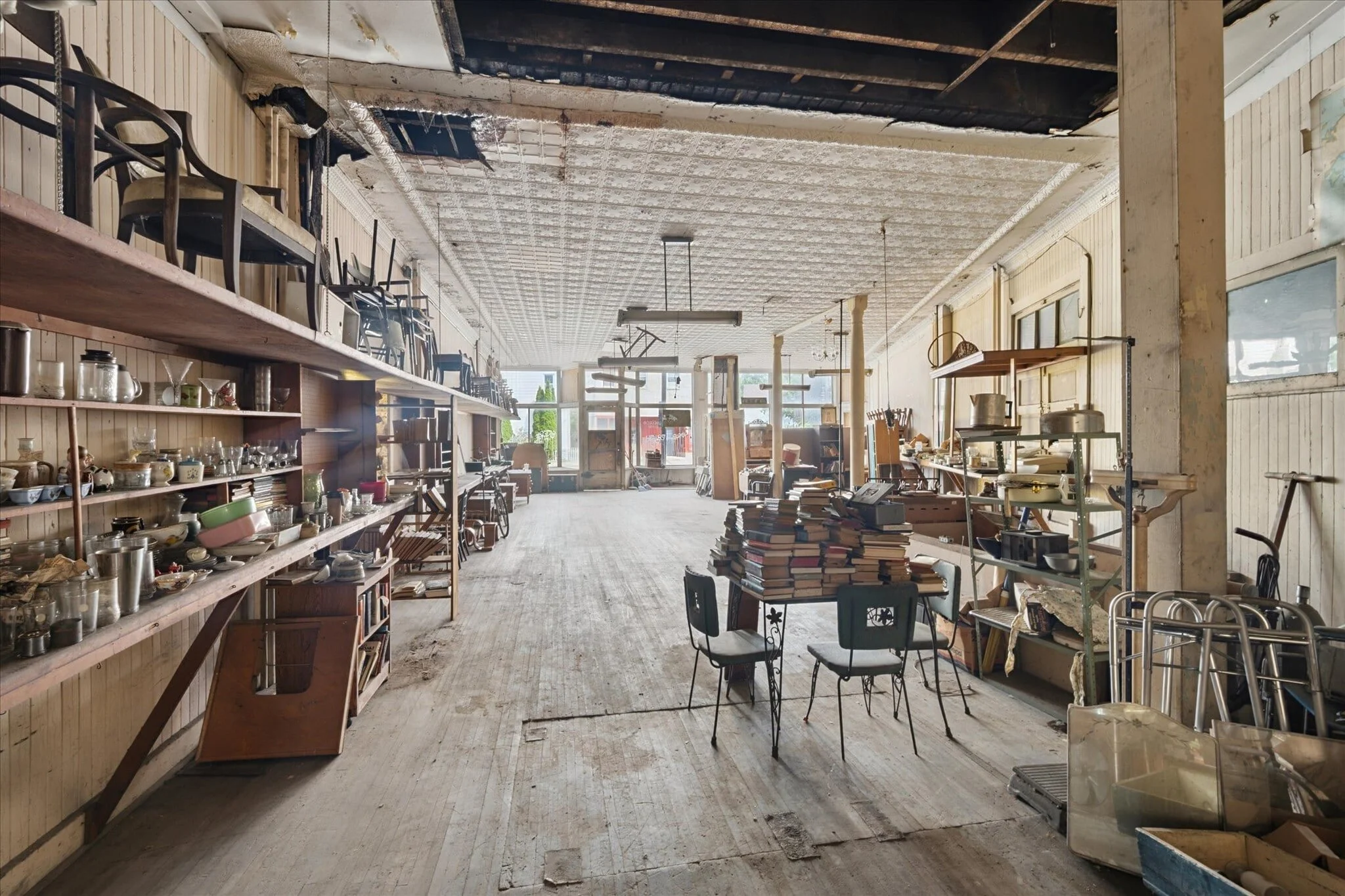Empty thrift store or warehouse with shelves of miscellaneous items, a pile of books on a table in the center, chairs stacked on the left, and sunlight coming through large windows.