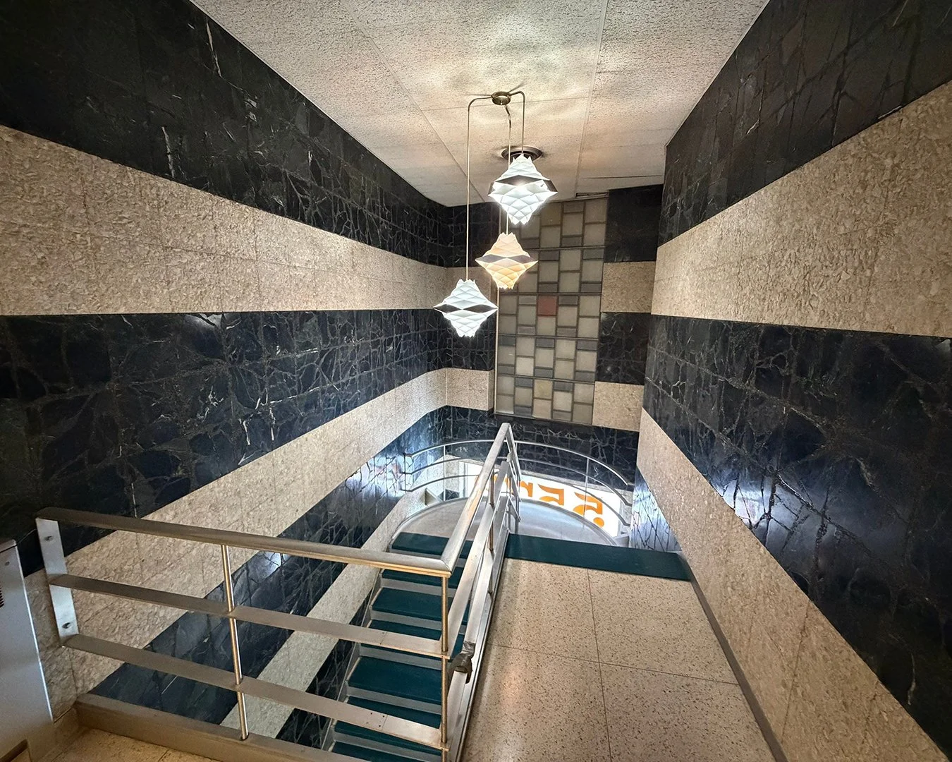 Interior view of a stairway in a building with black and beige striped walls, hanging pendant lights, a metal handrail, and a tiled section at the bottom.
