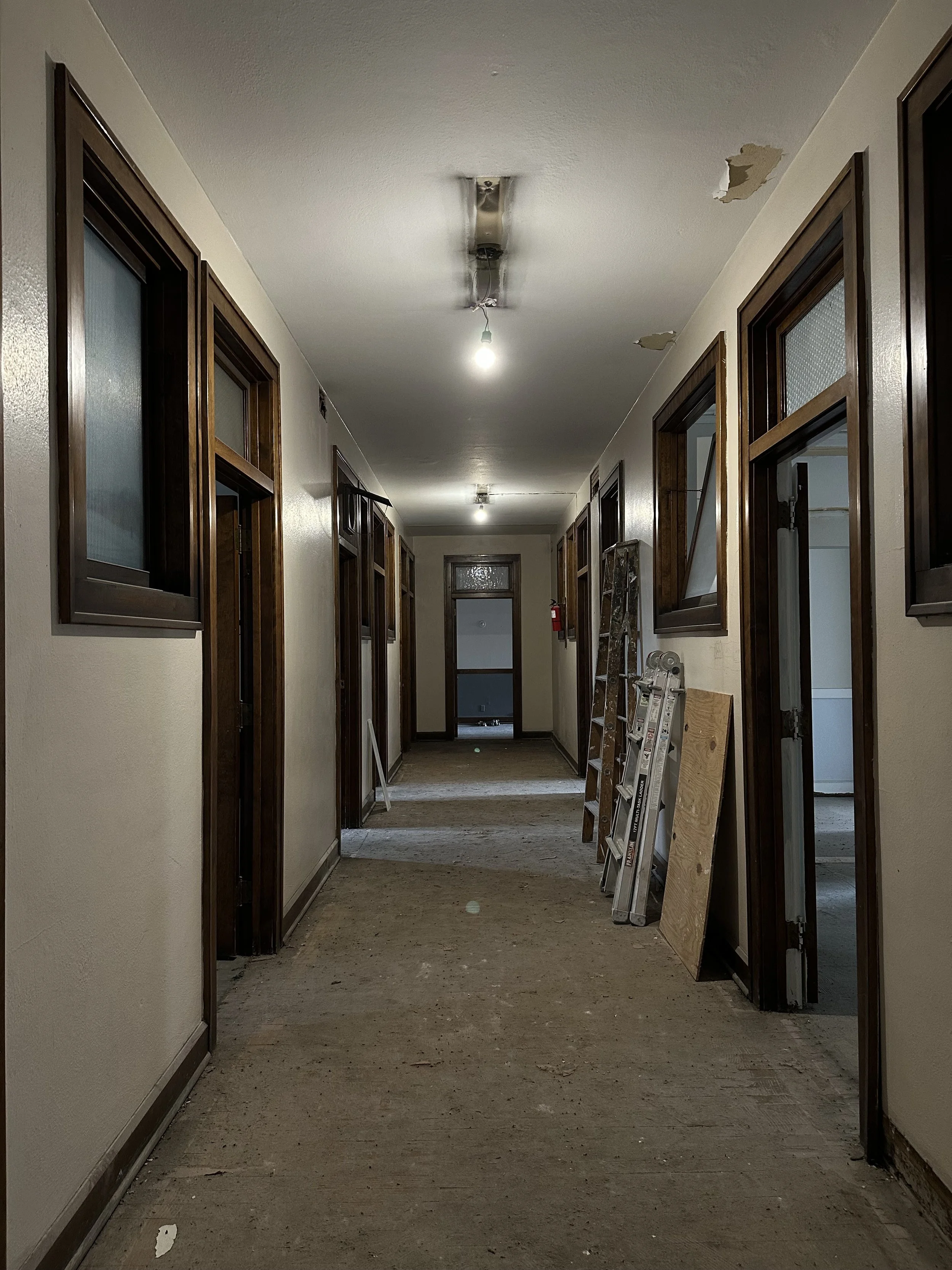 Underground corridor under renovation with wooden framed windows on both sides, unfinished floor, exposed ceiling with missing light fixtures, ladder and construction materials.