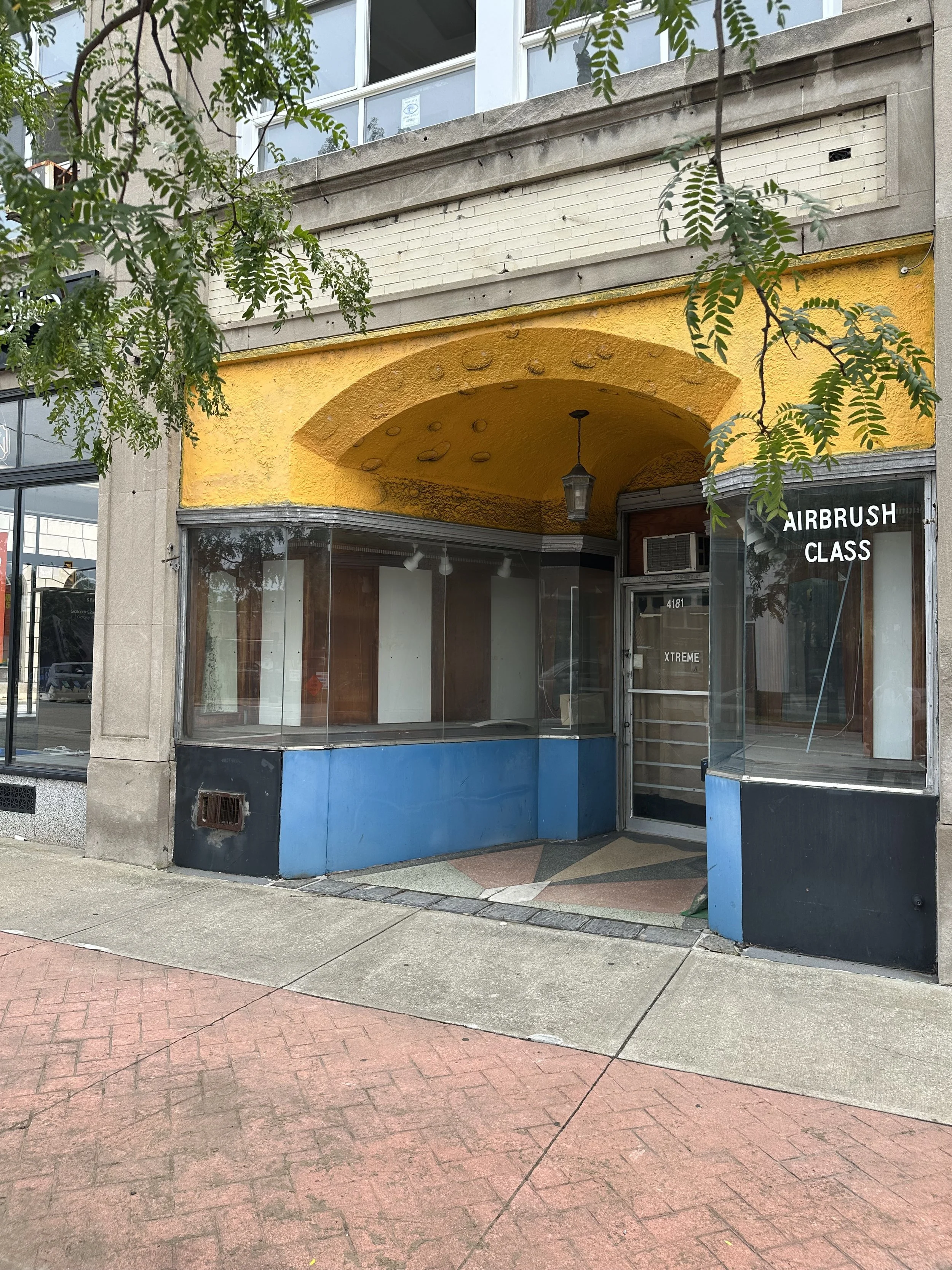 Empty storefront with large windows and an archway with a yellow textured ceiling, framed by blue and gray walls, on a city sidewalk with concrete and brick pavement, partially obscured by tree leaves.