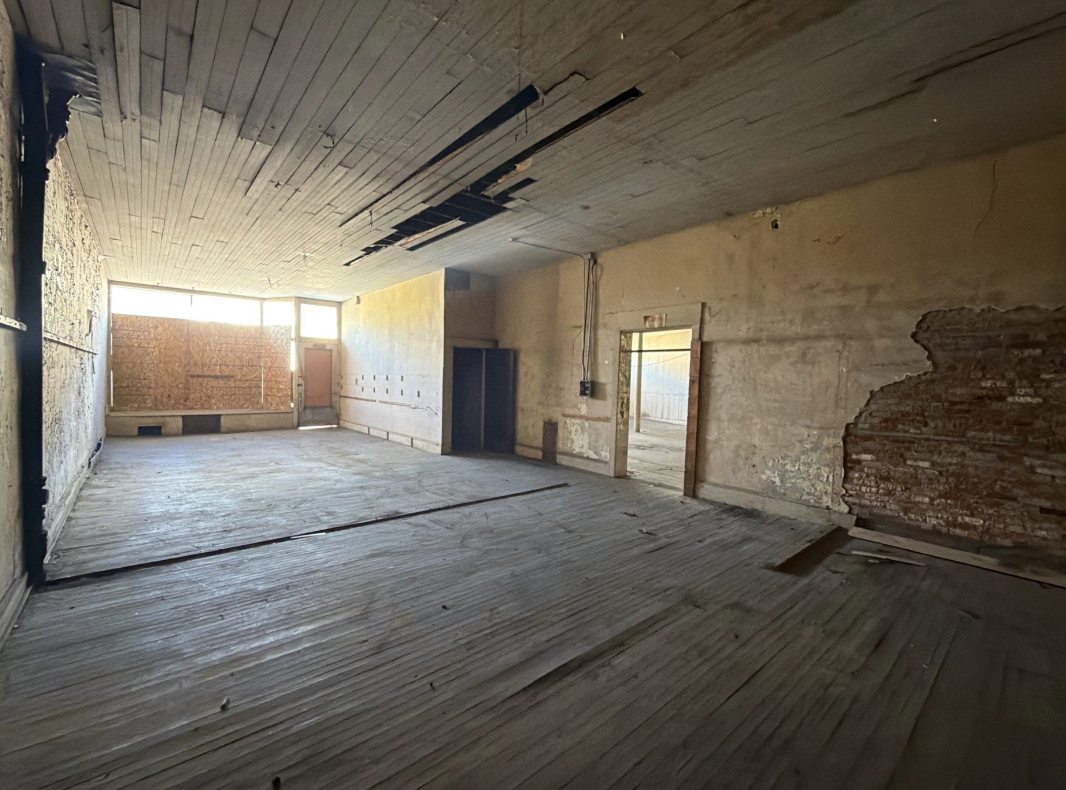 An empty, unfinished room with exposed wooden flooring, unpainted walls with visible damage and patchwork, a large window covered with cardboard, and a ceiling with peeling panels and visible wiring.