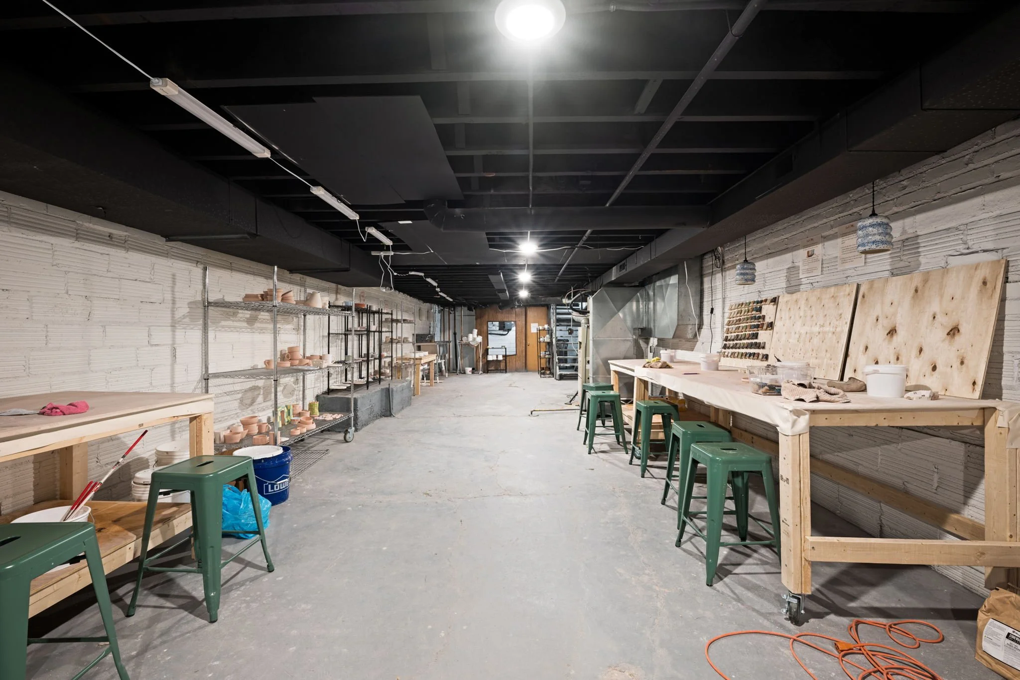 Empty pottery studio with worktables, chairs, and shelves holding pottery, in an industrial setting with exposed ceiling beams and bright overhead lighting.