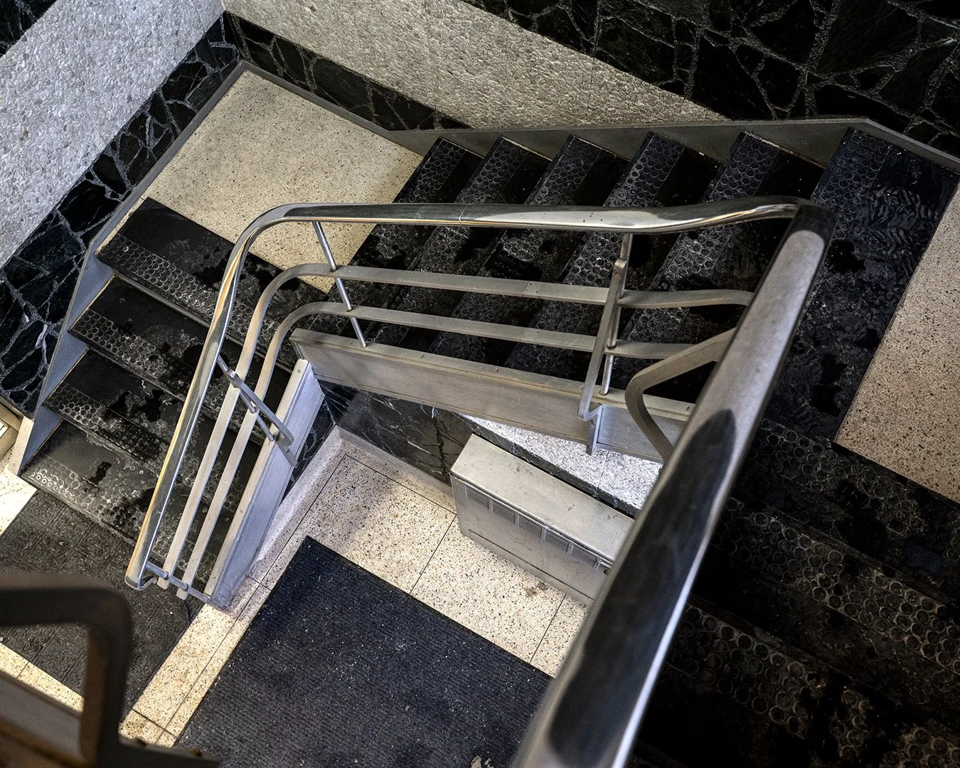 A top view of a staircase with black and beige speckled tiles, black rubber mats, and a silver metal handrail.