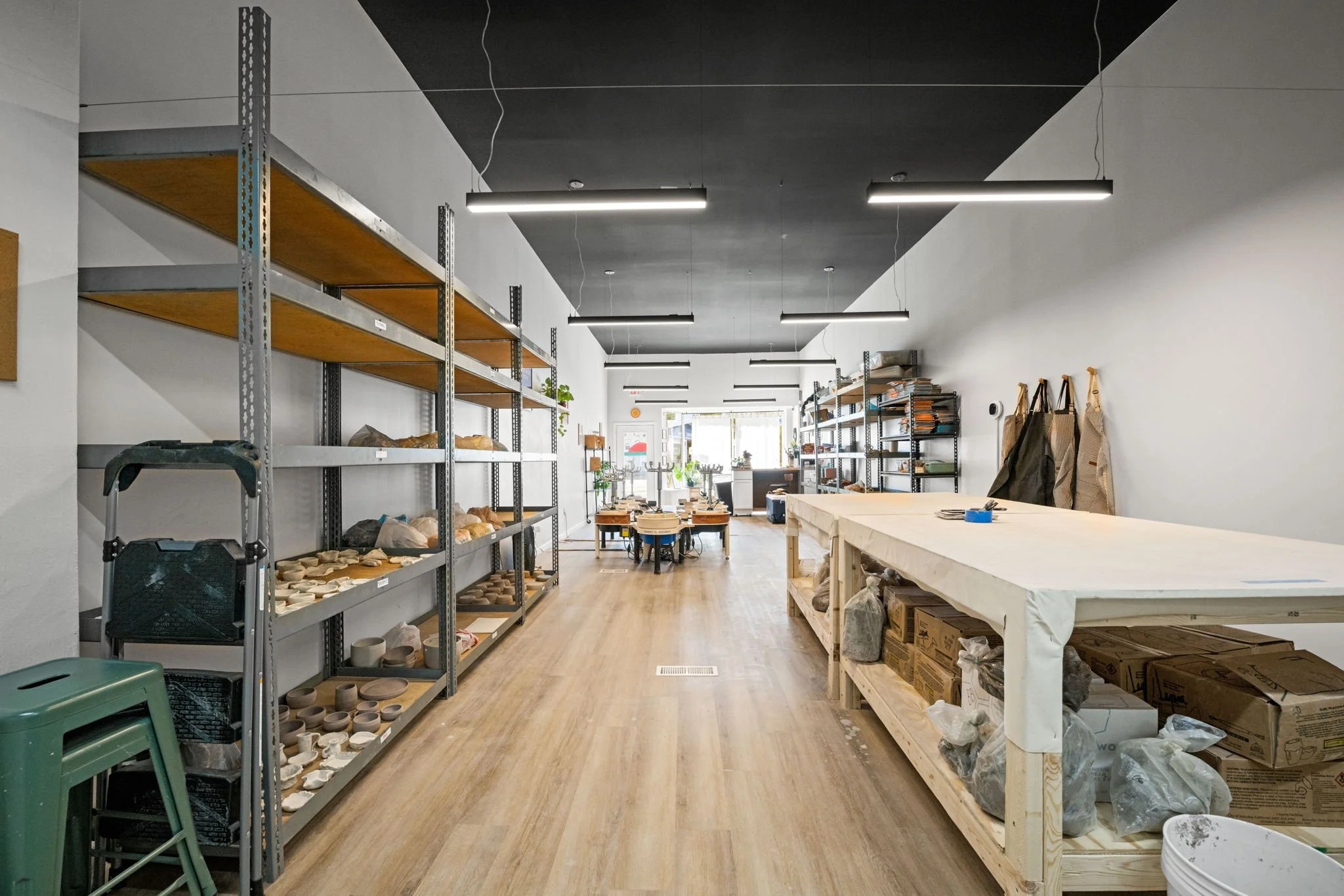 Interior of a ceramics studio with metal and wooden shelves holding pottery pieces and supplies, a large work table with bags and boxes, and a dining area with a table and chairs in the background, illuminated by overhead lighting.