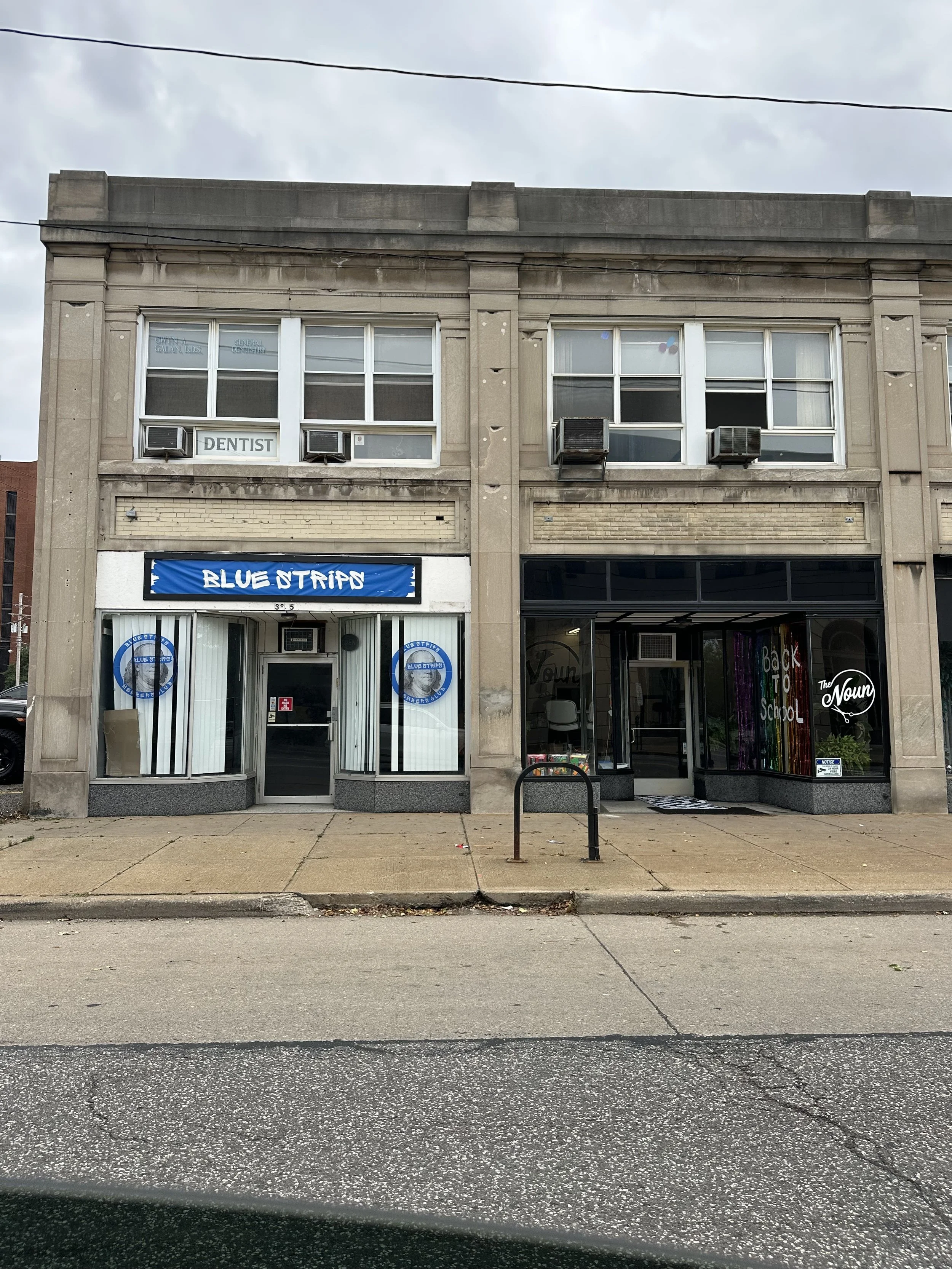 A two-story building with a dental office on the second floor, indicated by a sign saying 'DENTIST' in the window. The lower level has a storefront with a blue sign that reads 'BLUE STRIP' and another shop with a rainbow-colored sign. The sidewalk ha