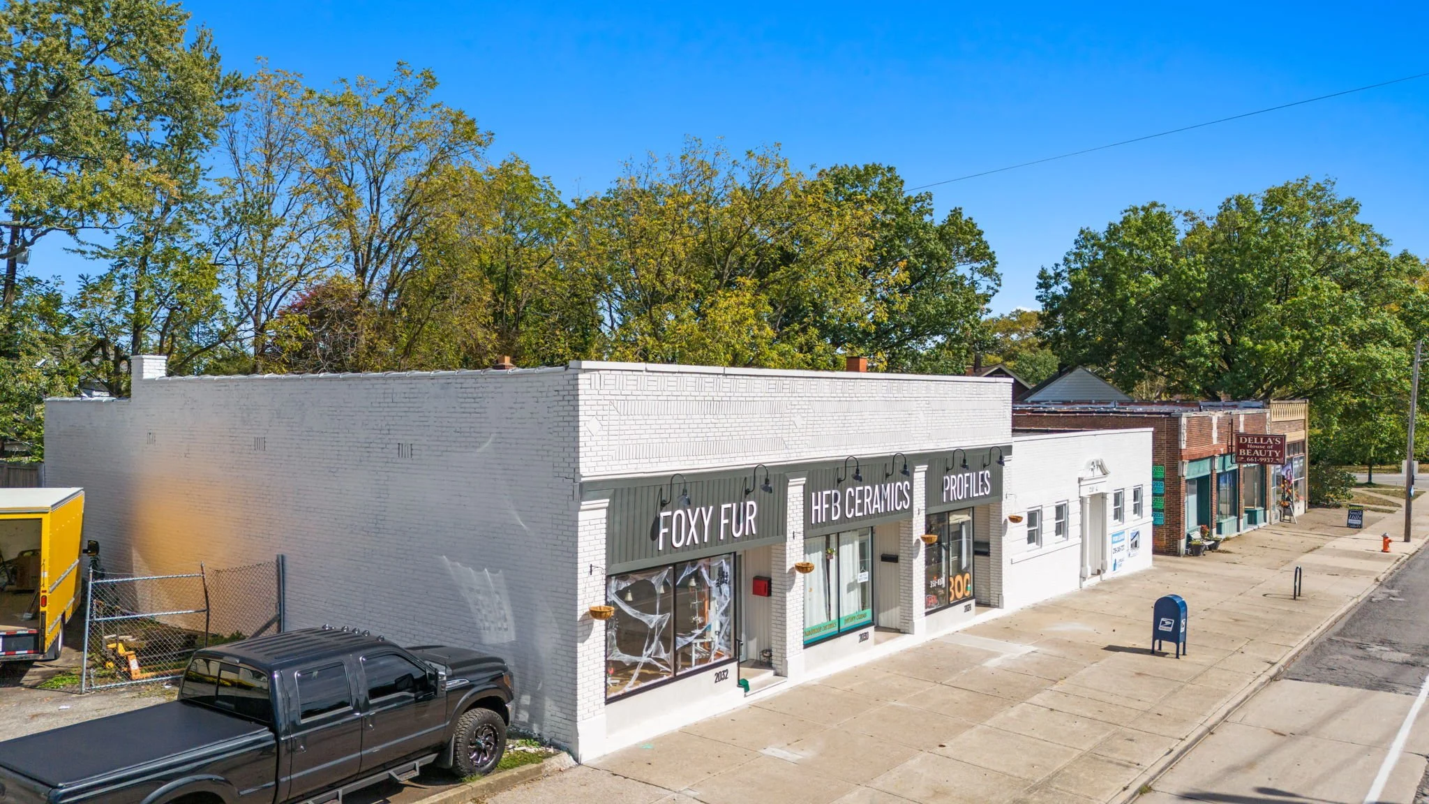 A row of small storefronts on a street with a blue sky, trees in the background, and parked vehicles including a black pickup truck in front, featuring signs for Foxy Fur, HFB Ceramics, Profiles, and Dellas Beauty.