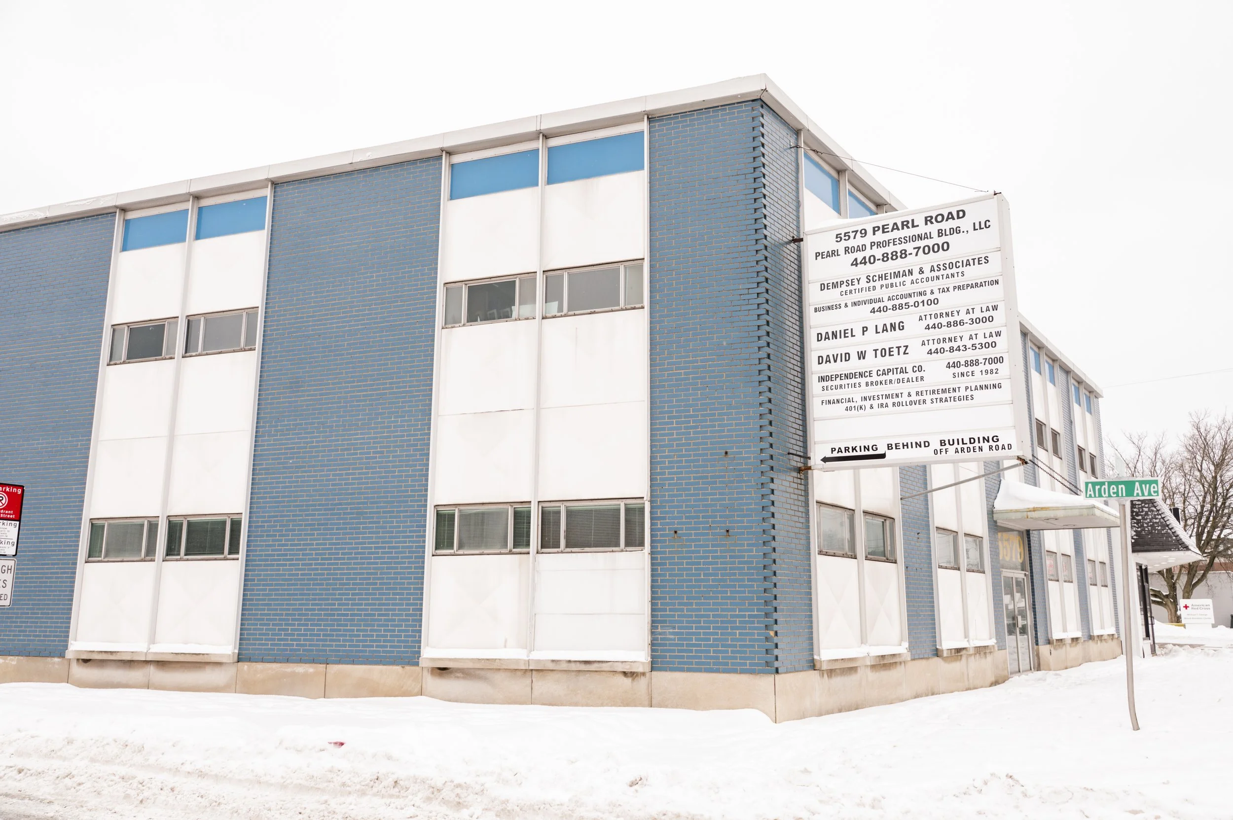 Multi-story building with blue brick exterior, white window panels, and a large white sign with business information, located on a snowy street corner with street sign reading 'Arden Ave'.