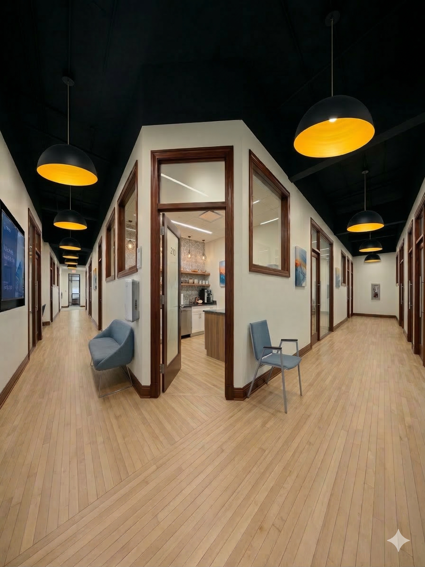 Office or commercial building hallway with a wooden floor, cream-colored walls, and multiple doors with wooden trims. There are chairs along the walls, modern hanging lights with yellow interiors, and some artwork on the walls.