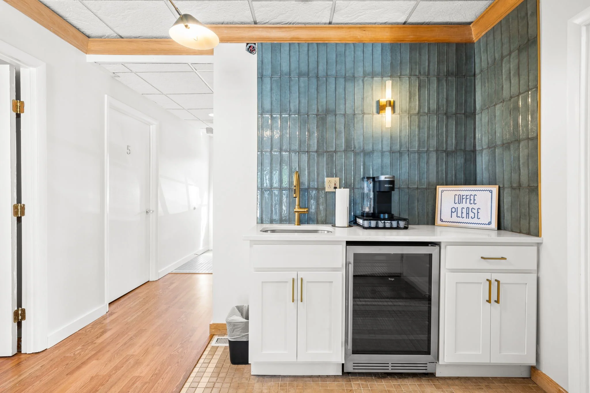 A small kitchenette with a white countertop, a gold faucet, and a coffee machine, set against a blue-tiled wall with a wall light, with a sign that says 'Coffee Please.'