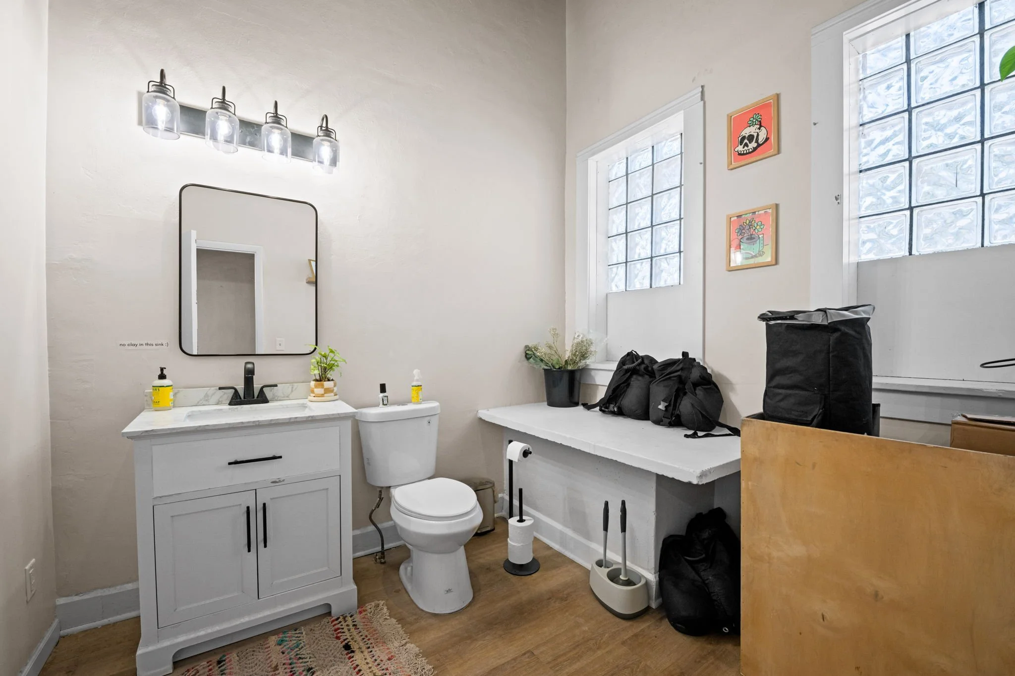 Bathroom with white vanity, mirror, toilet, and two windows. Decor includes pictures with skull and plant motifs, and various items on a countertop and shelf.