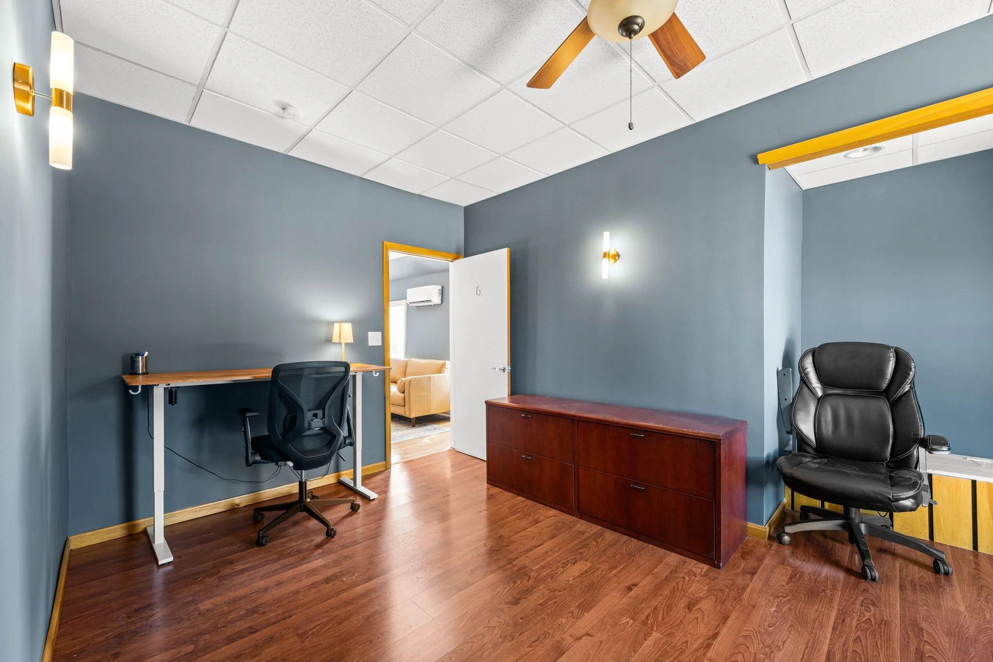 An office with blue walls and wooden flooring, featuring a desk with an office chair, a wooden cabinet, and a black leather office chair. There are wall-mounted lights, a ceiling fan, and an open door leading to a room with a yellow couch.