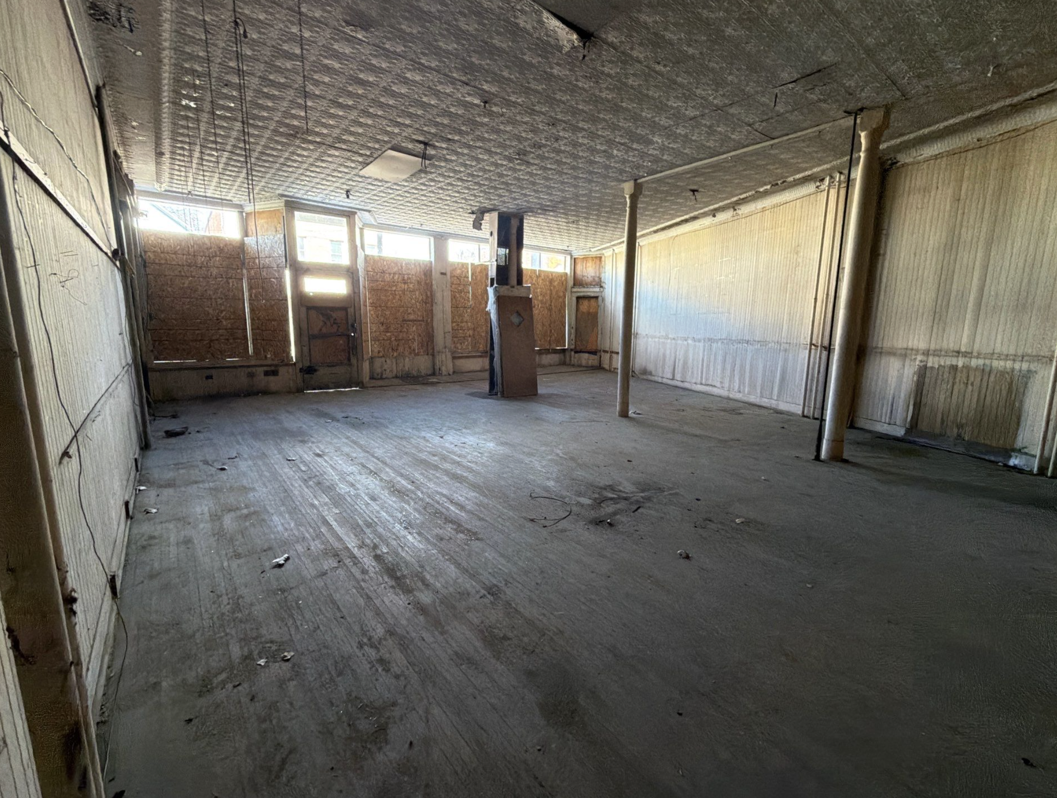 Empty, unfinished room with plywood covering windows, exposed ceiling, and support poles in the center.