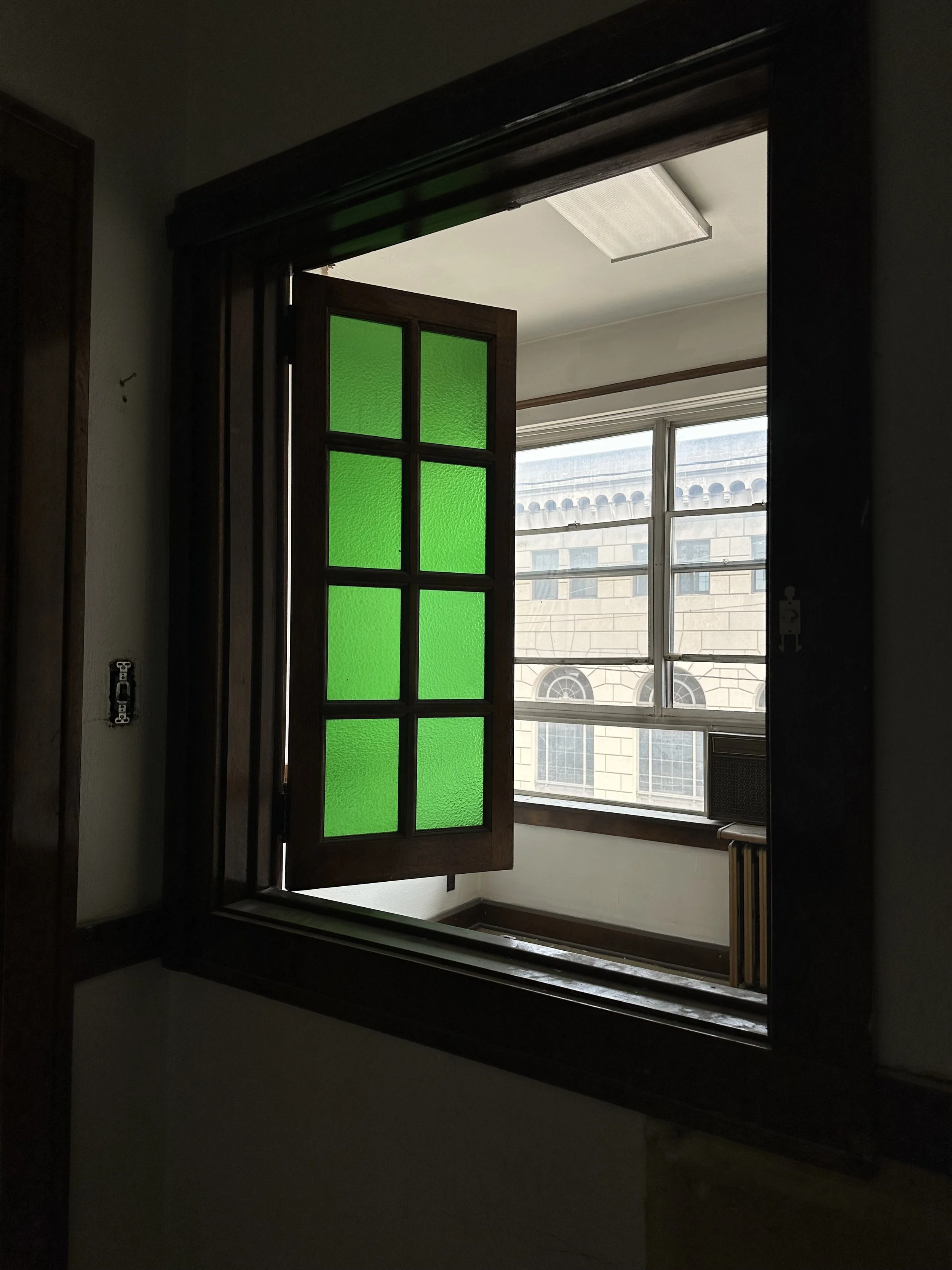 An empty room with a large window and a partially opened wooden window with green stained glass panels.