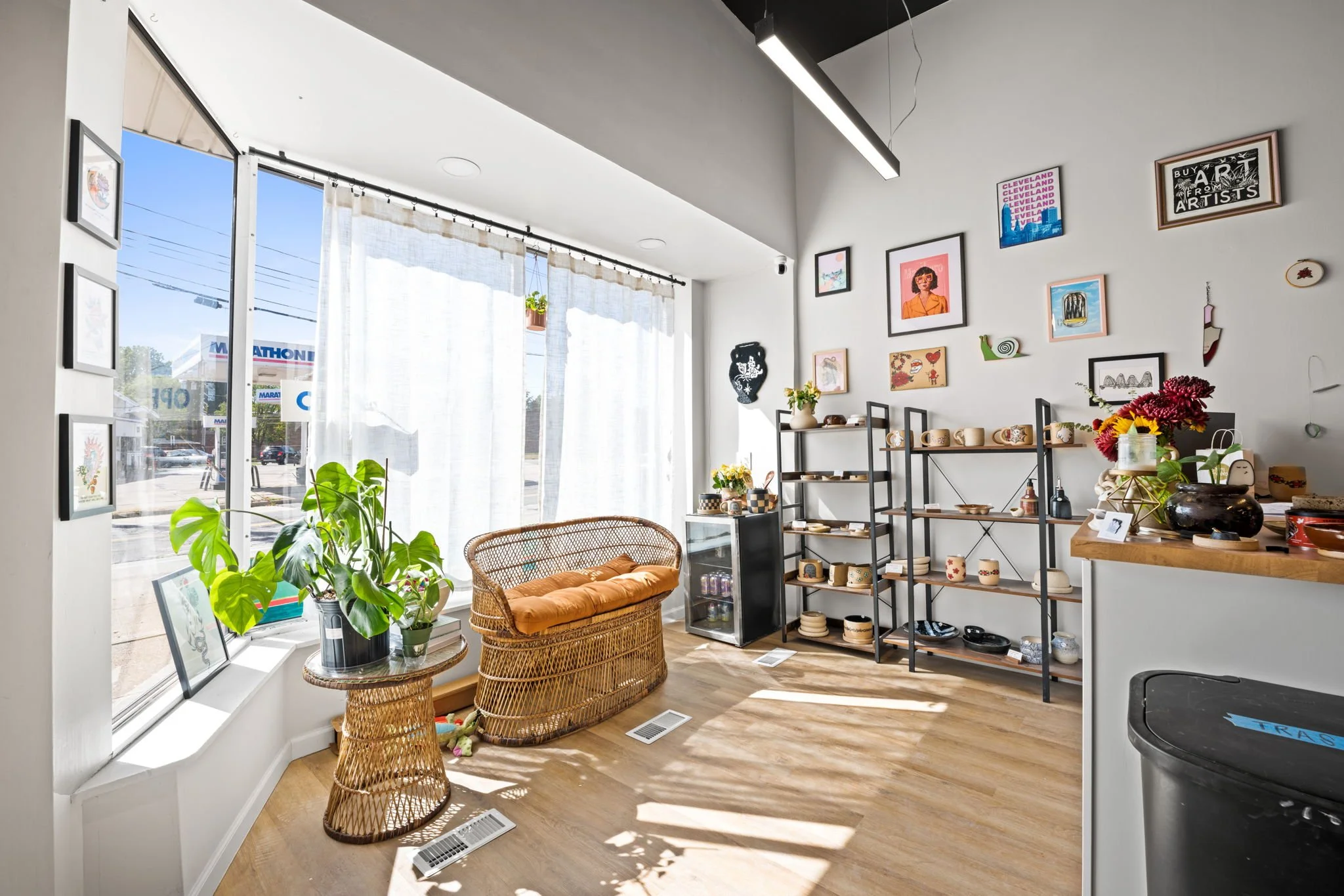 Bright interior space with large front window draped with white curtains, wooden floors, black metal shelving units displaying ceramics and decorative items, framed artwork and posters on white walls, wicker loveseat with orange cushion, potted plant