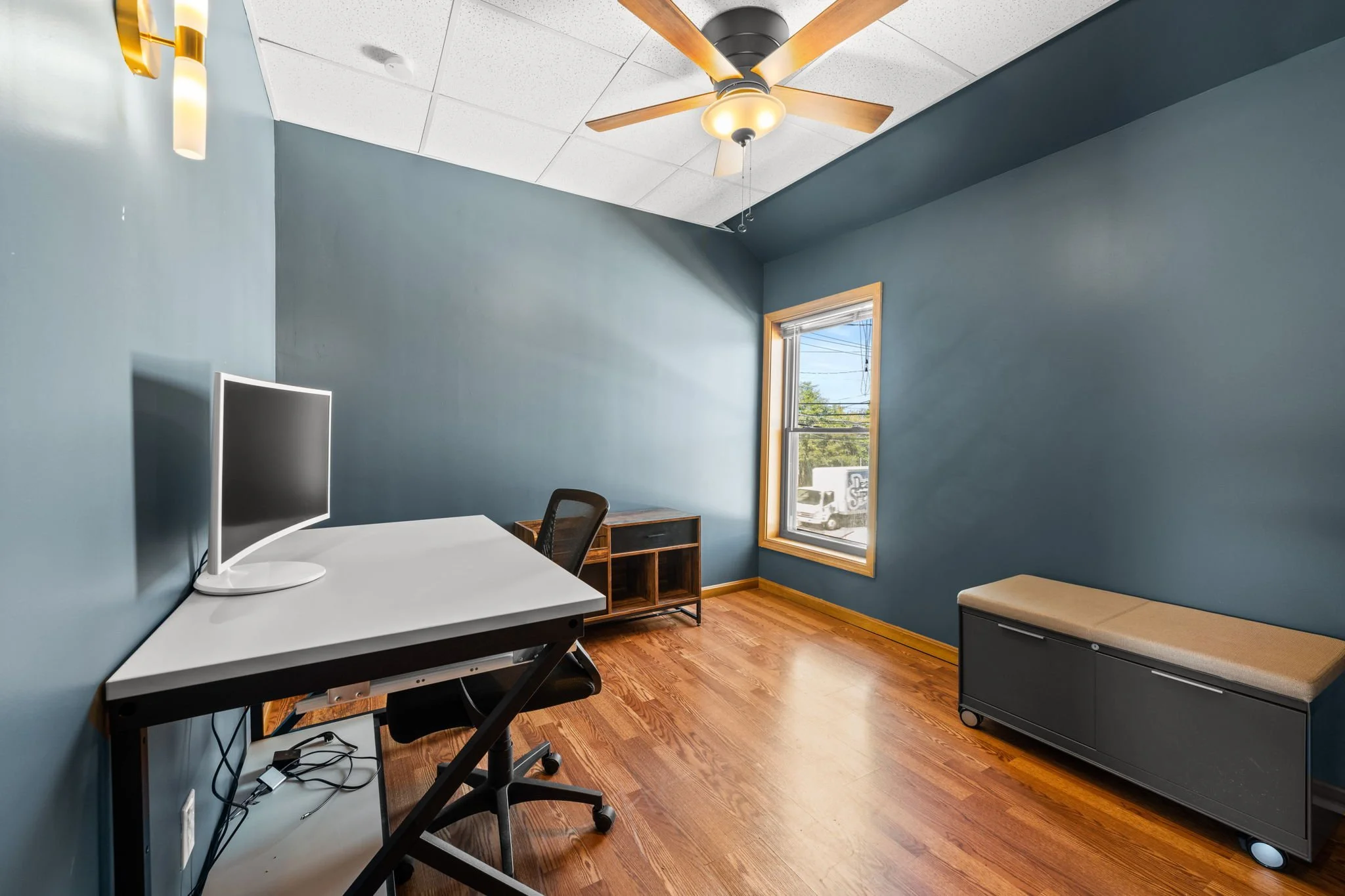 An empty home office with blue walls, a window with blinds, a ceiling fan, a white desk with a computer monitor, a black office chair, a small wooden desk, and a black storage bench with a beige cushion.