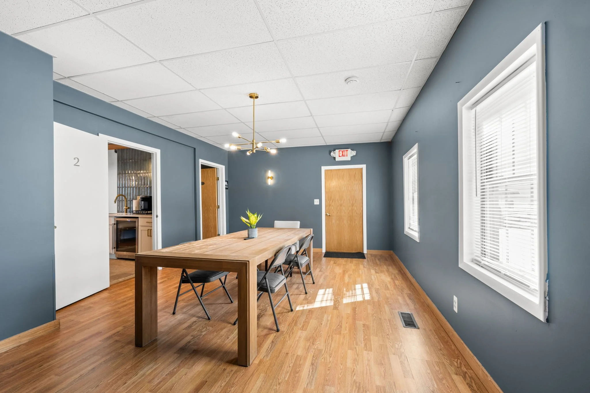 Empty dining room with blue walls, wooden floor, a long wooden table, and four chairs. Bright windows with white blinds, a modern chandelier, and an exit door.