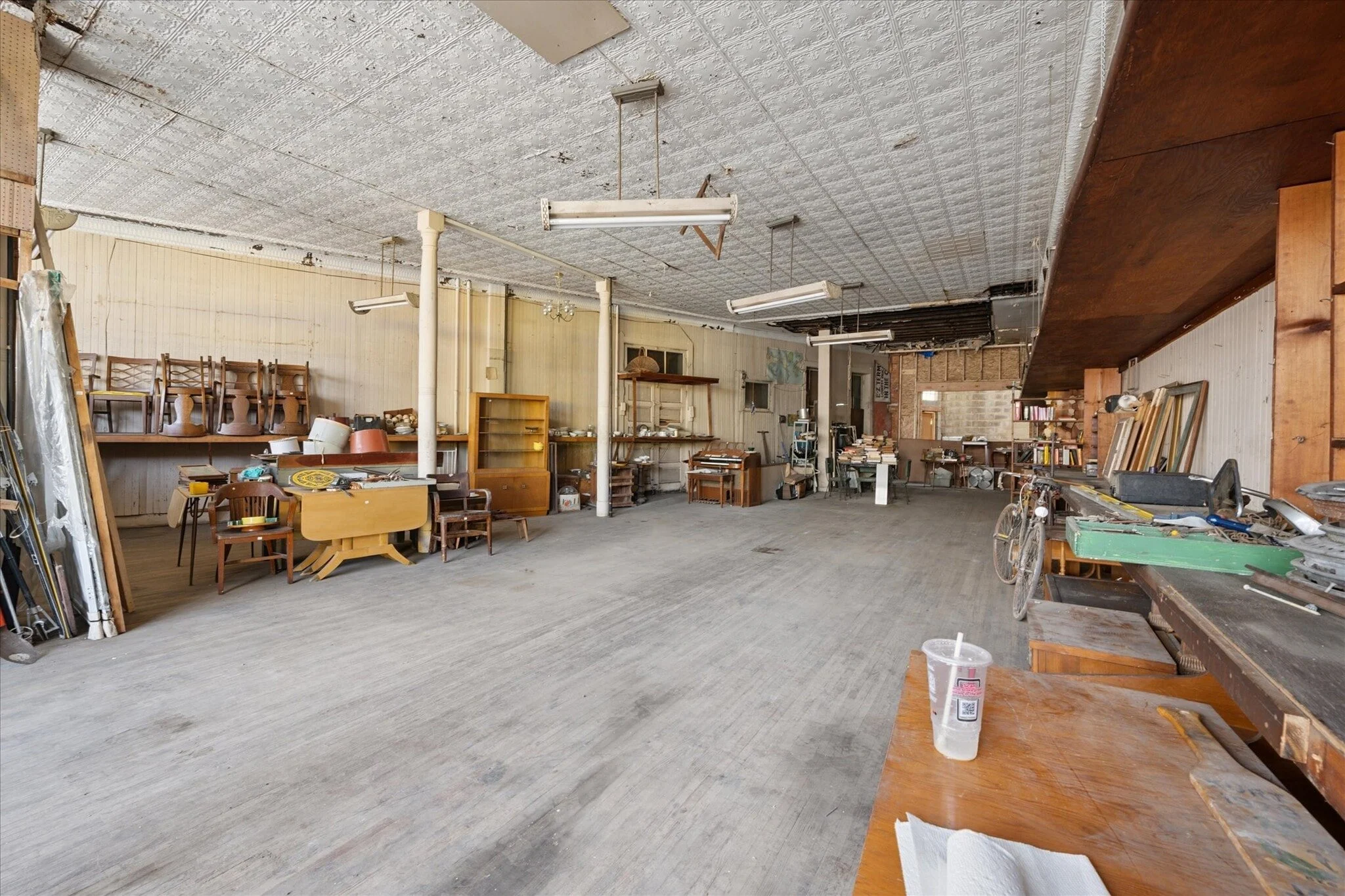 Interior view of a spacious, cluttered room with various furniture, tools, and shelves. The ceiling has exposed tiles and hanging lights. The floor is wood, and the walls are partially paneled and partly unfinished.
