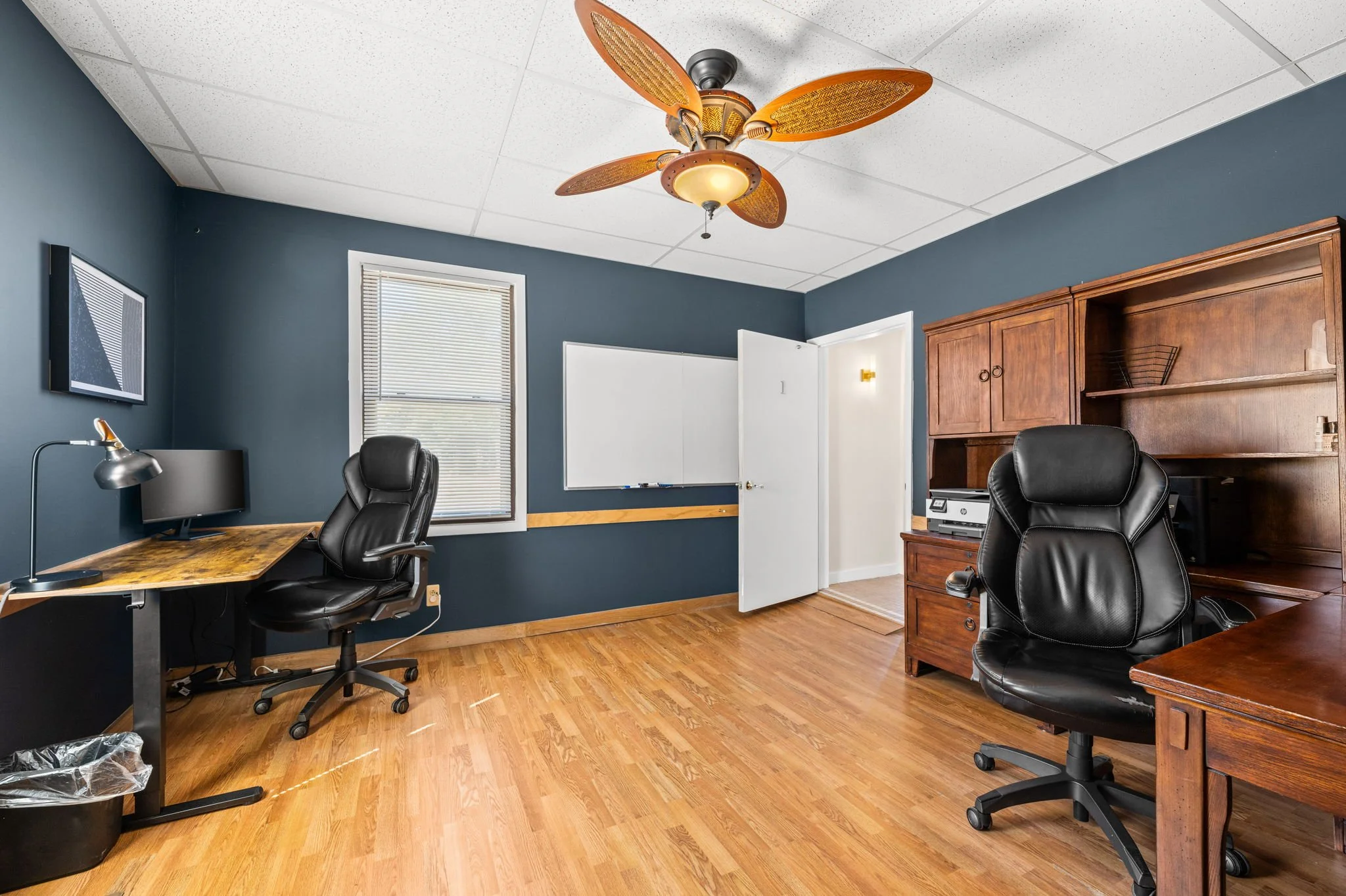 Office room with wooden flooring, dark blue walls, ceiling fan with palm leaf design, two black office chairs, a wooden desk, file cabinet, wall-mounted monitor, window with blinds, whiteboard, and open door.