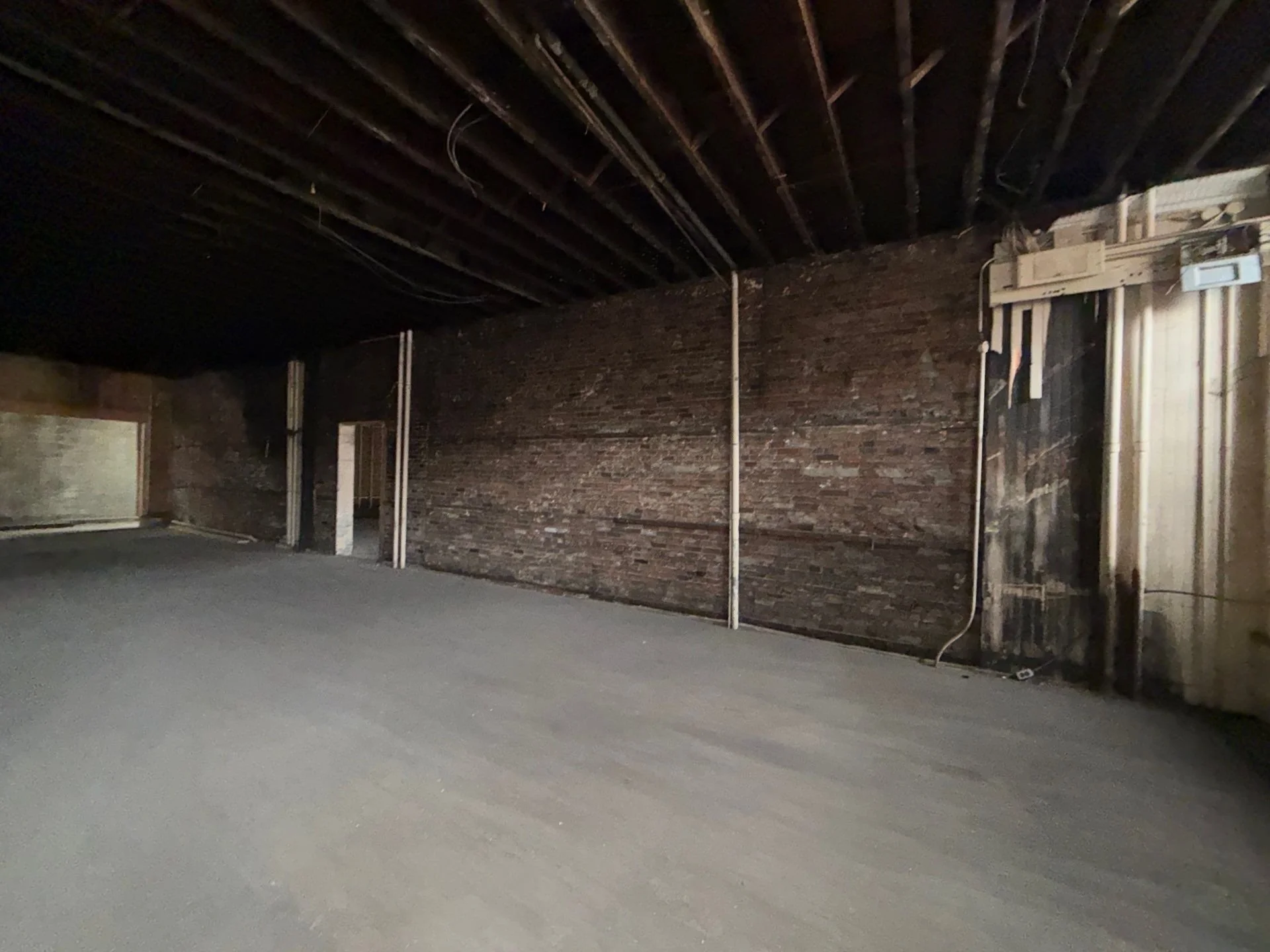 Interior of an unfinished room with exposed brick wall, black ceiling beams, and exposed wiring. The floor appears to be concrete.