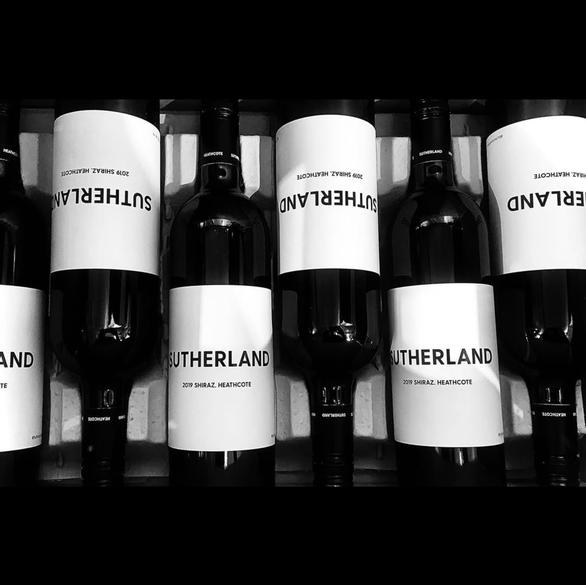 Multiple bottles of Sutherland 2019 Shiraz wine with white labels stacked upside down on a shelf.