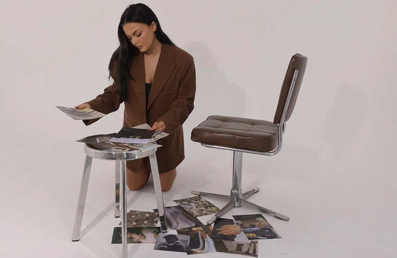 A woman with long dark hair kneels on the floor, wearing a brown blazer over black clothing, surrounded by scattered photographs and looking at one photograph. A small round table with more photographs is in front of her, and a brown office chair with a metal base is positioned nearby against a plain white background.