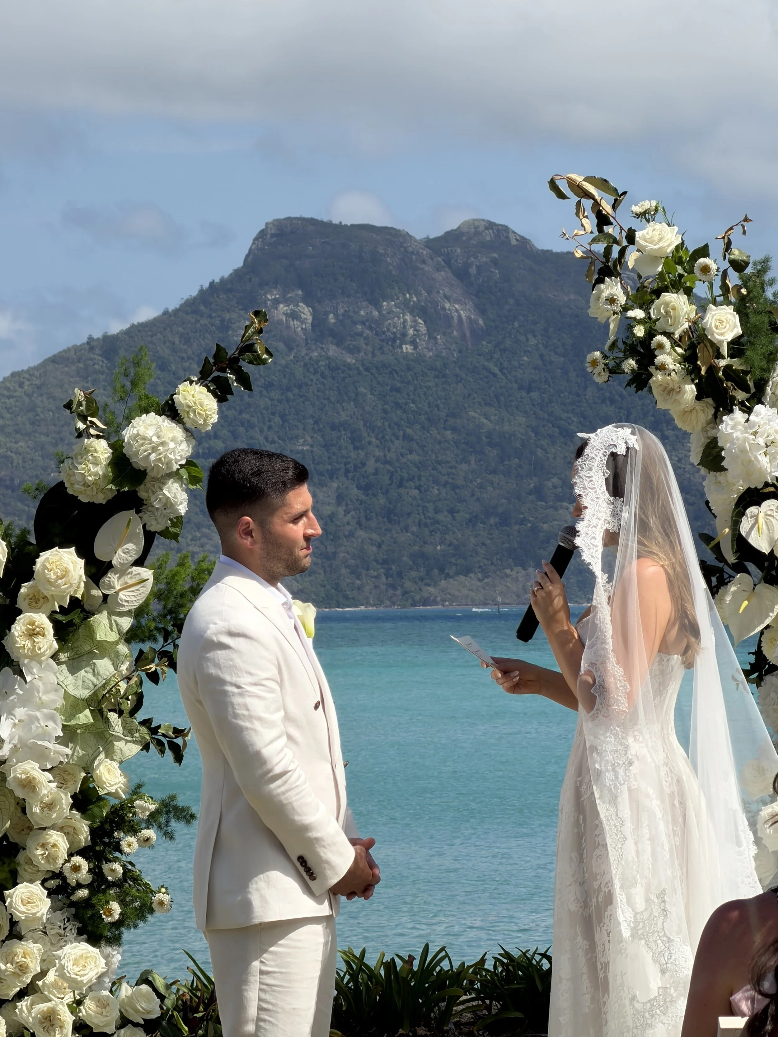 A bride and groom are having a wedding ceremony outdoors near a body of water with mountains in the background. The bride is reading her vows into a microphone, and they are standing under a floral arch decorated with white roses and greenery.