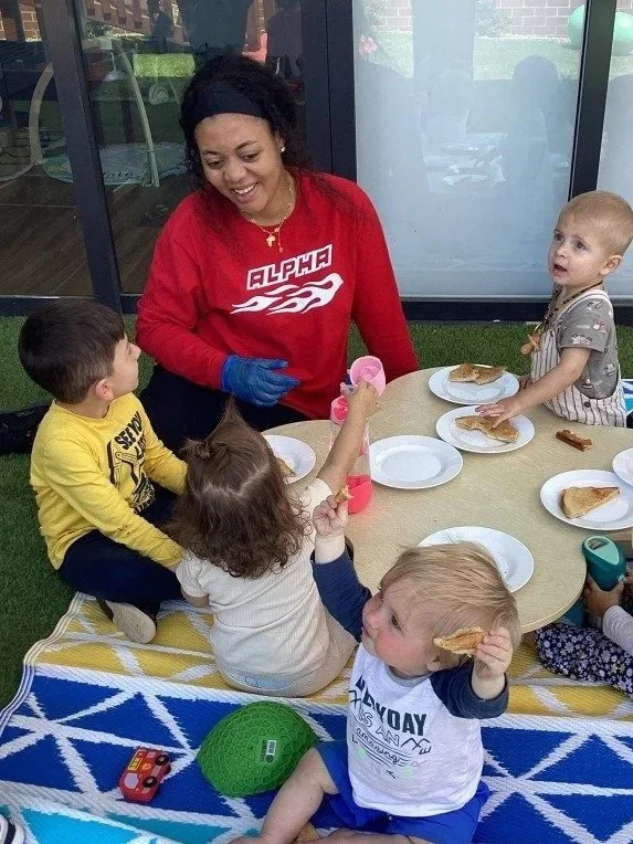 A young boy looking at himself in a mirror attached to a wooden play structure in a playroom, with another play area visible in the background.
