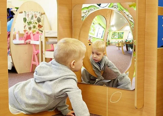 A young boy looking at himself in a mirror attached to a wooden play structure in a playroom, with another play area visible in the background.