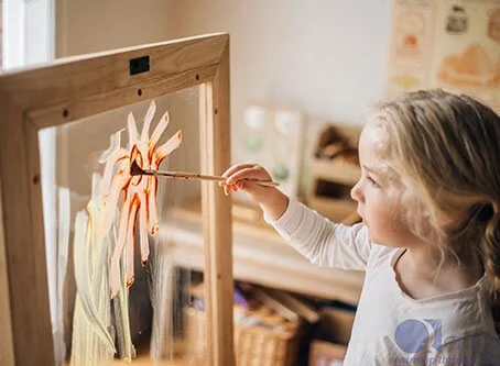A young girl is painting with a brush on a glass easel, creating a colorful abstract design.