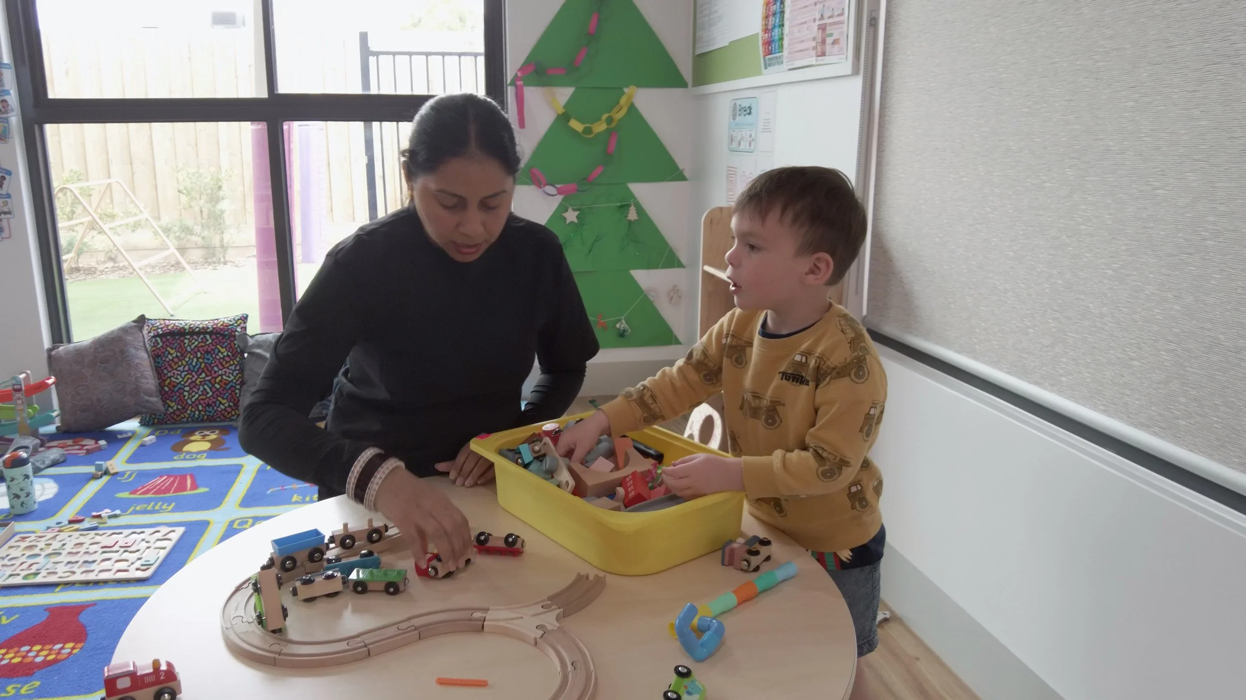 A young girl sitting cross-legged on the floor playing with a wooden fairy tale treehouse toy surrounded by small pinecones, tree-shaped figures, and other miniature wooden animals in a cozy room.