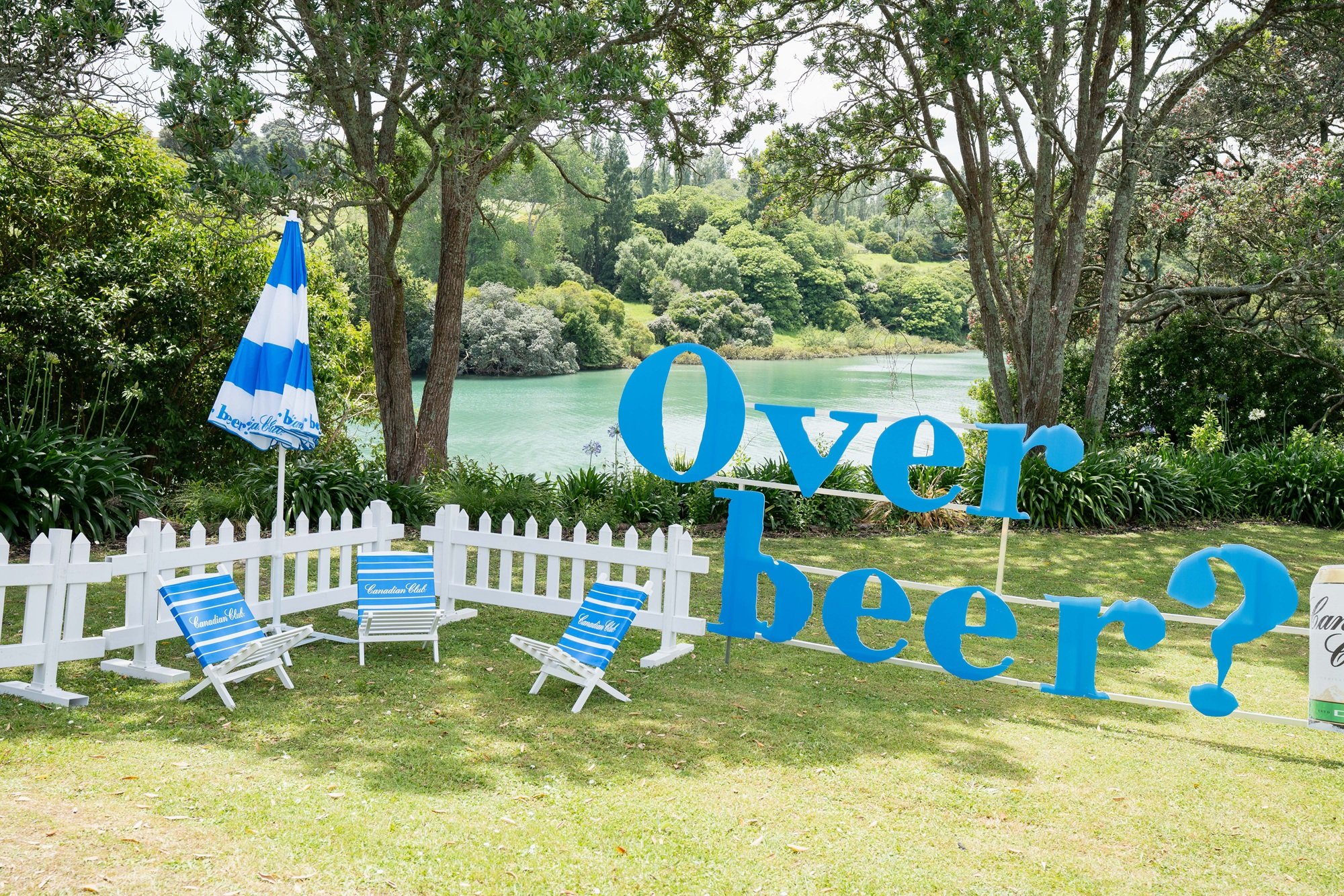 Area with a white picket fence, three blue and white striped chairs labeled 'Canadian Club', and a blue and white striped umbrella. Large blue letters spell out 'Over here?' near the water, with a scenic view of trees, bushes, and a lake in the background.