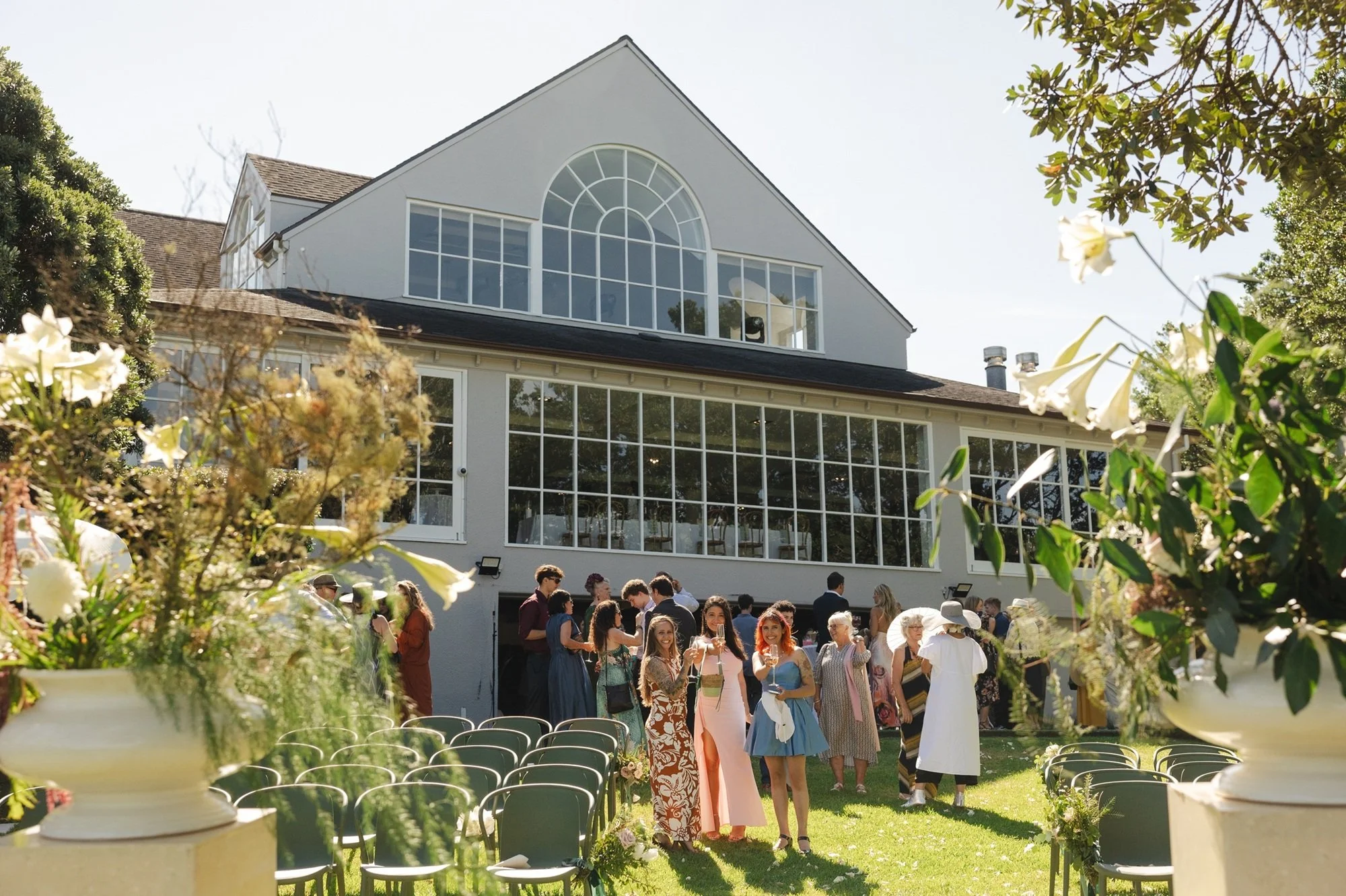 People gathered outside a large white house with many windows, decorated for a special event or celebration on a sunny day.