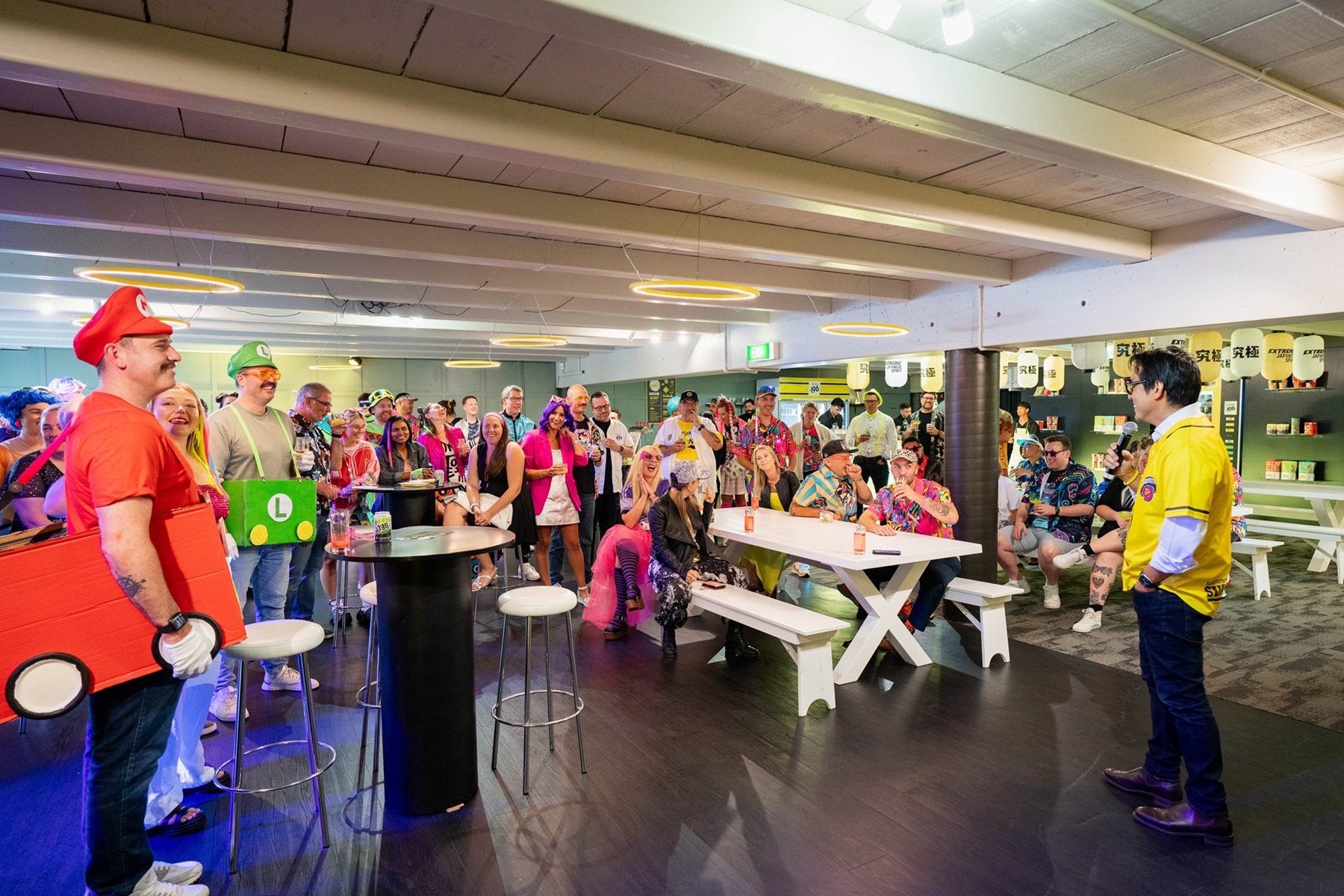 Group of people at a costume party or celebration, many dressed as characters from the Mario video game, listening to a man in a yellow jersey speaking into a microphone in a decorated indoor space.