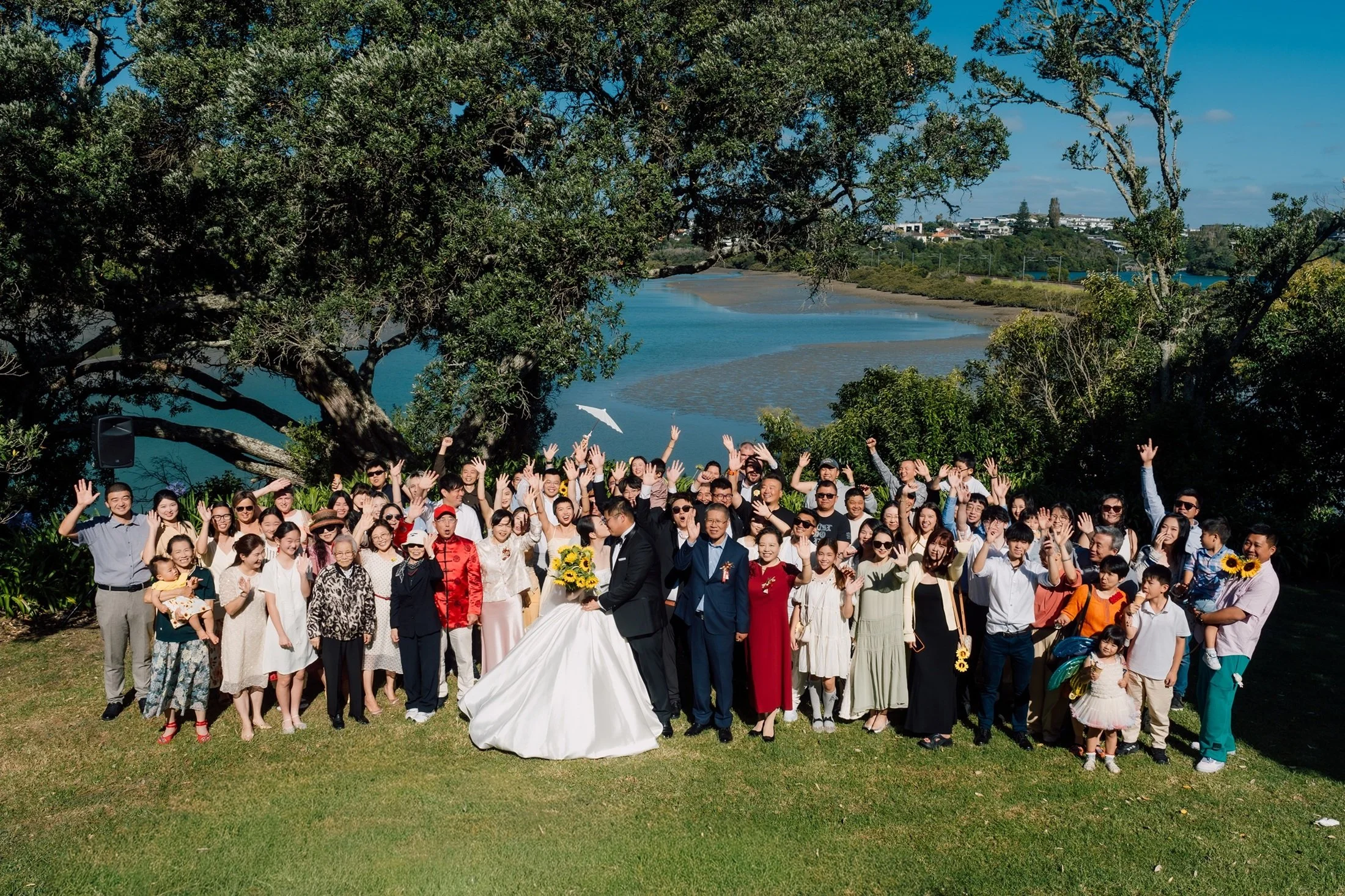 A large group of people celebrating a wedding outdoors by a river on a sunny day, with many smiling and waving at the camera. The bride in a white gown and the groom in a dark suit are in front, surrounded by family and friends, some holding sunflowers and children.