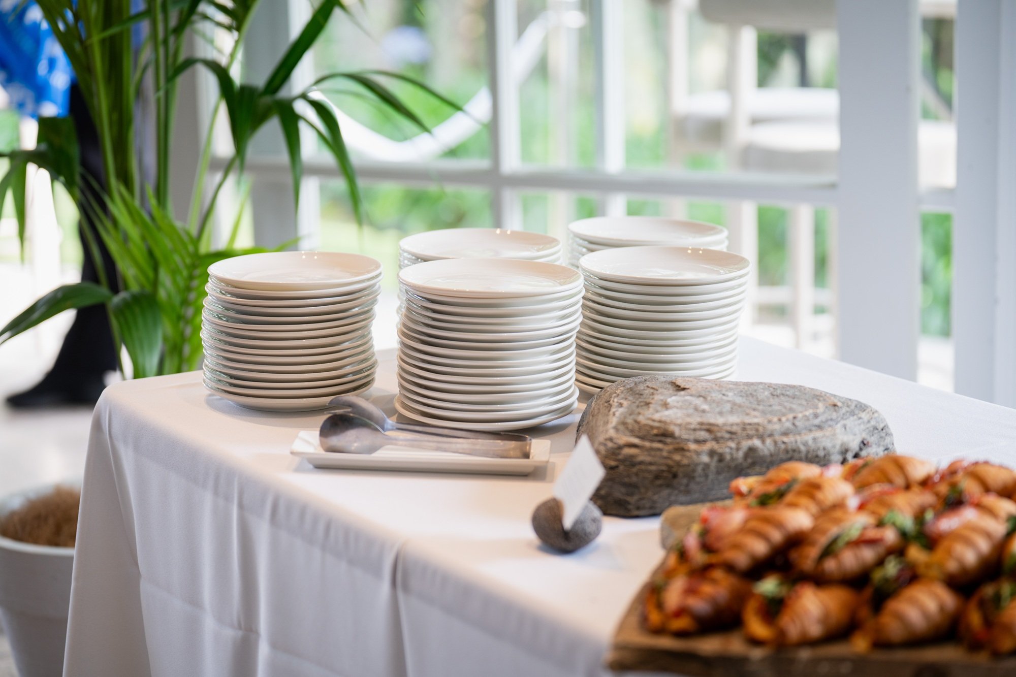 Stacks of white plates on a table set for a buffet, with a large rock and some pastry in the foreground, near a window with natural light.