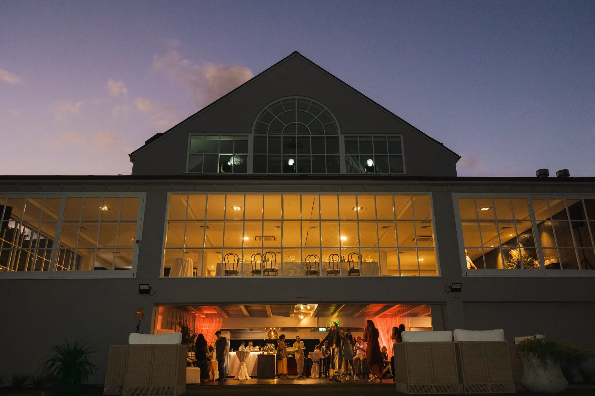 People socializing at an evening event inside and outside a multi-story house with large glass windows and warm interior lighting.